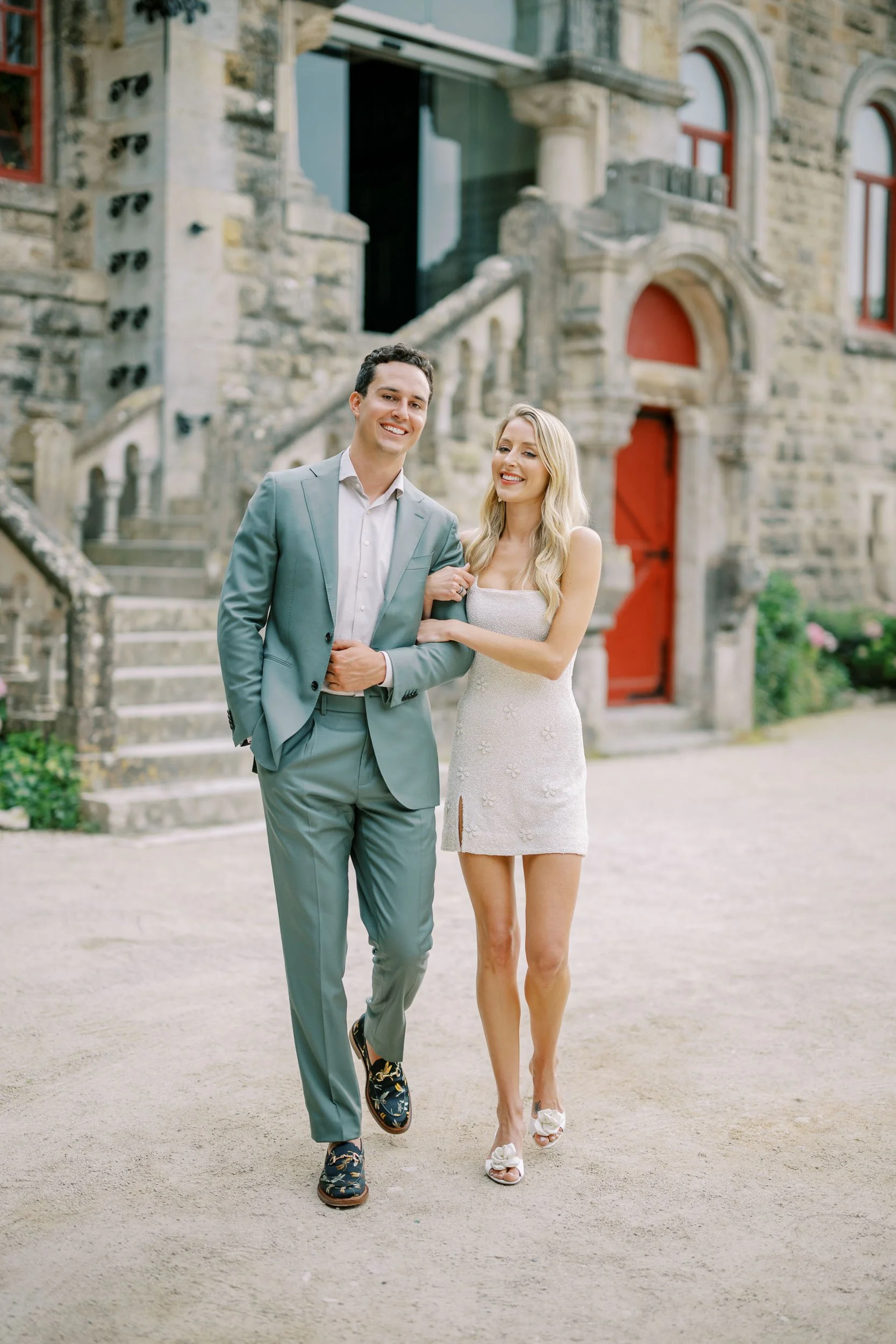 A happy couple walking arm-in-arm outside a historic stone building with red doors and stairs, dressed in formal attire at Forte da Cruz in Cascais, Portugal