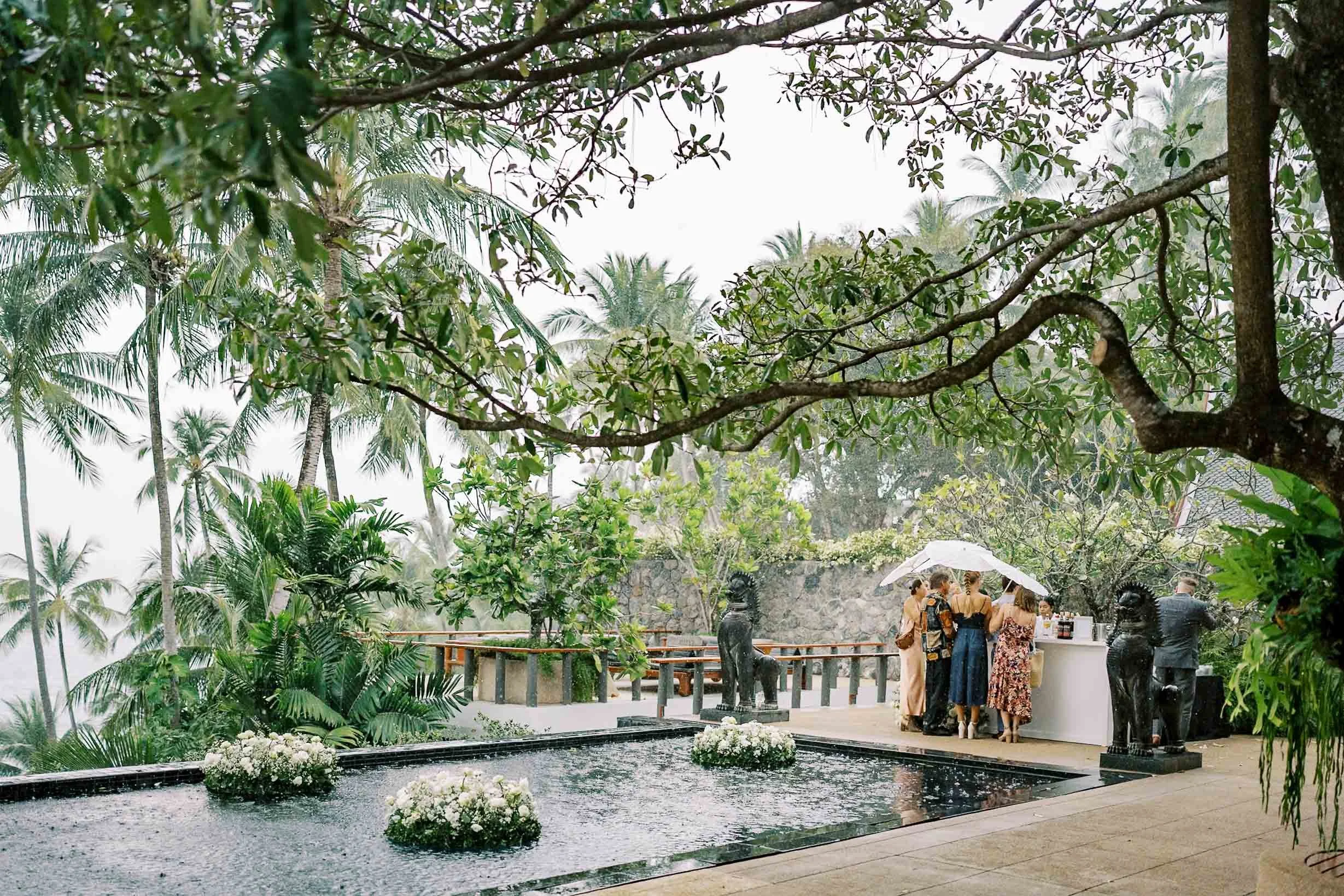 Rain falling by the pool as guests gather with umbrellas at Amanpuri in Phuket, Thailand