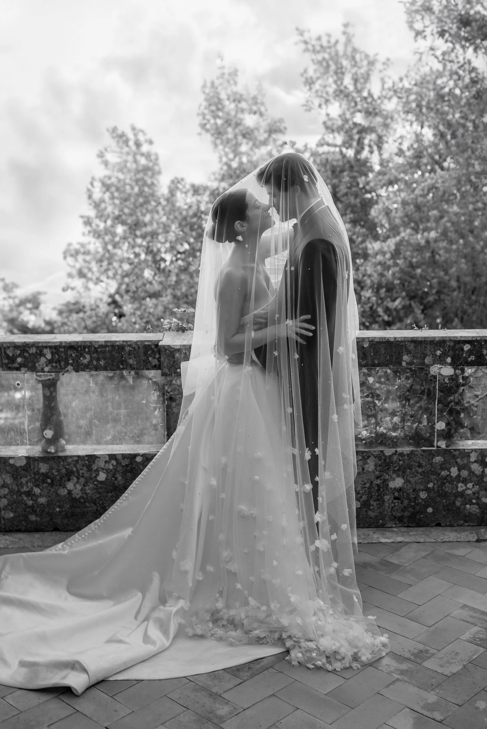 Black and white photo of a bride and groom standing close, kissing under a veil outdoors. The bride wears a strapless wedding gown with a long train and a veil with floral embellishments. The groom wears a dark suit. Trees and a cloudy sky are in the