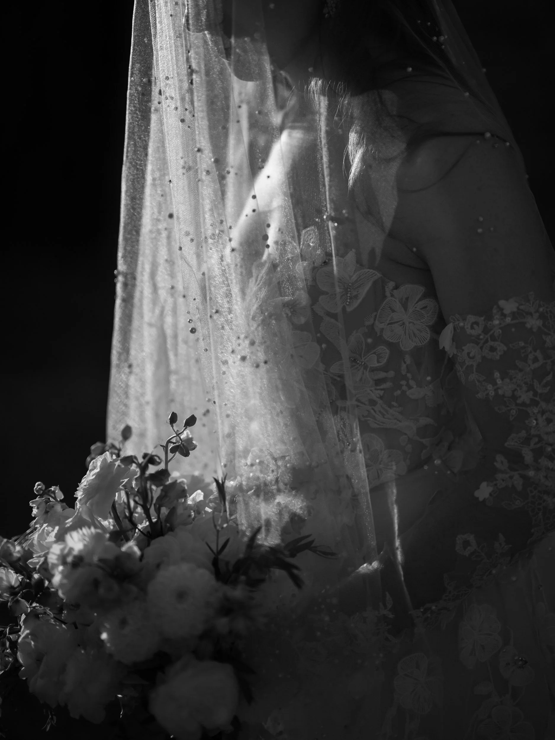 A bride in a lace dress holding a bouquet of flowers, partially obscured by a veil, in black and white.