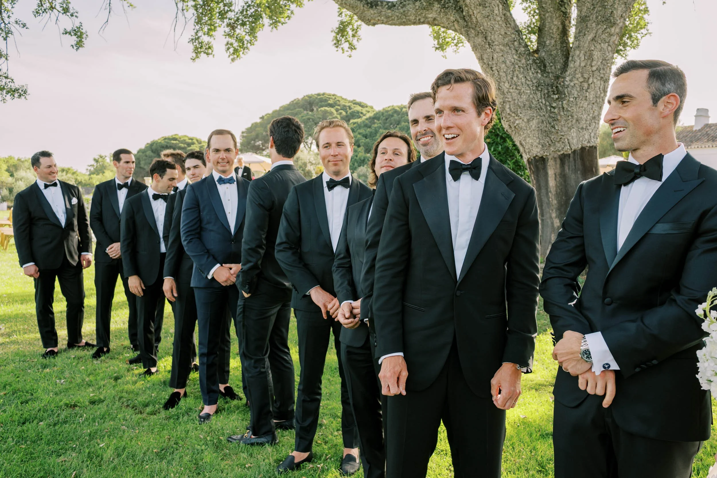 A group of groomsmen dressed in black tuxedos and bow ties standing outdoors in a line under a large tree, smiling and engaging in conversation during a formal outdoor event.