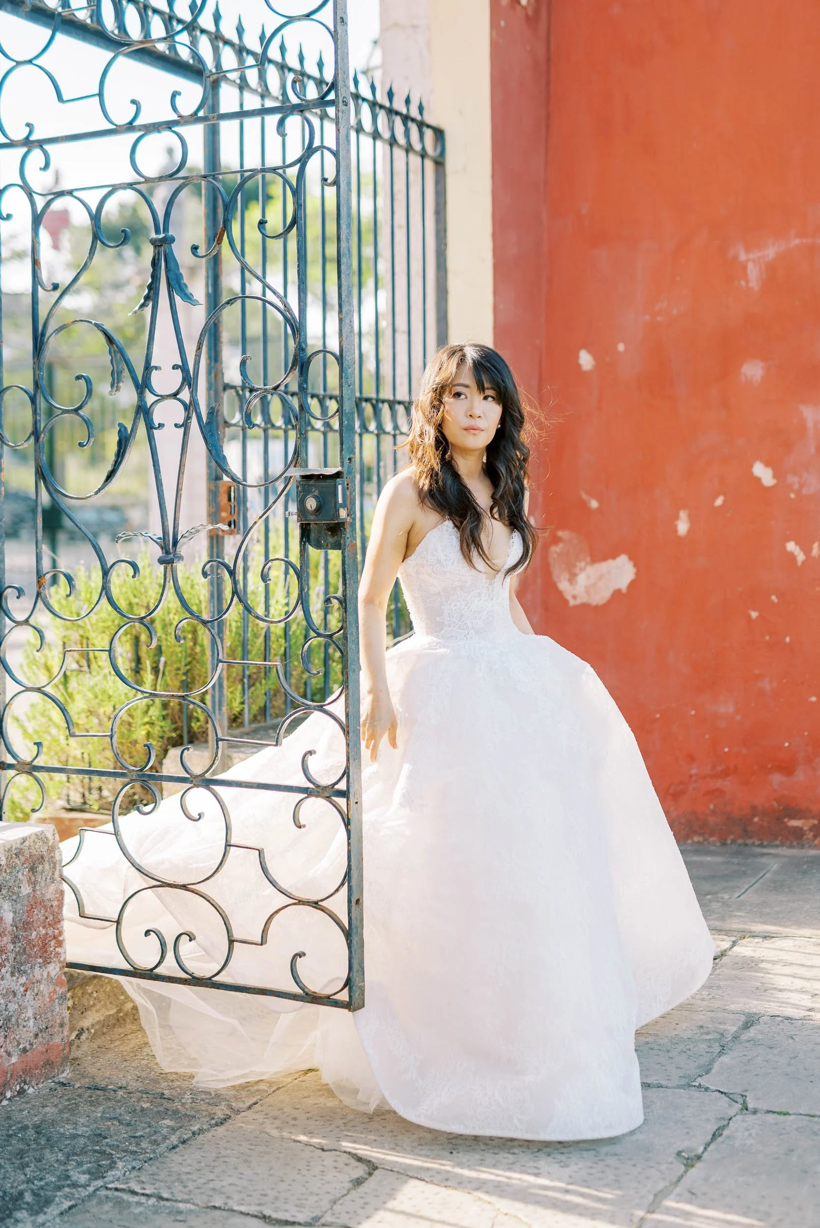 A bride in a wedding dress standing at an open wrought iron gate, outdoors during daytime.