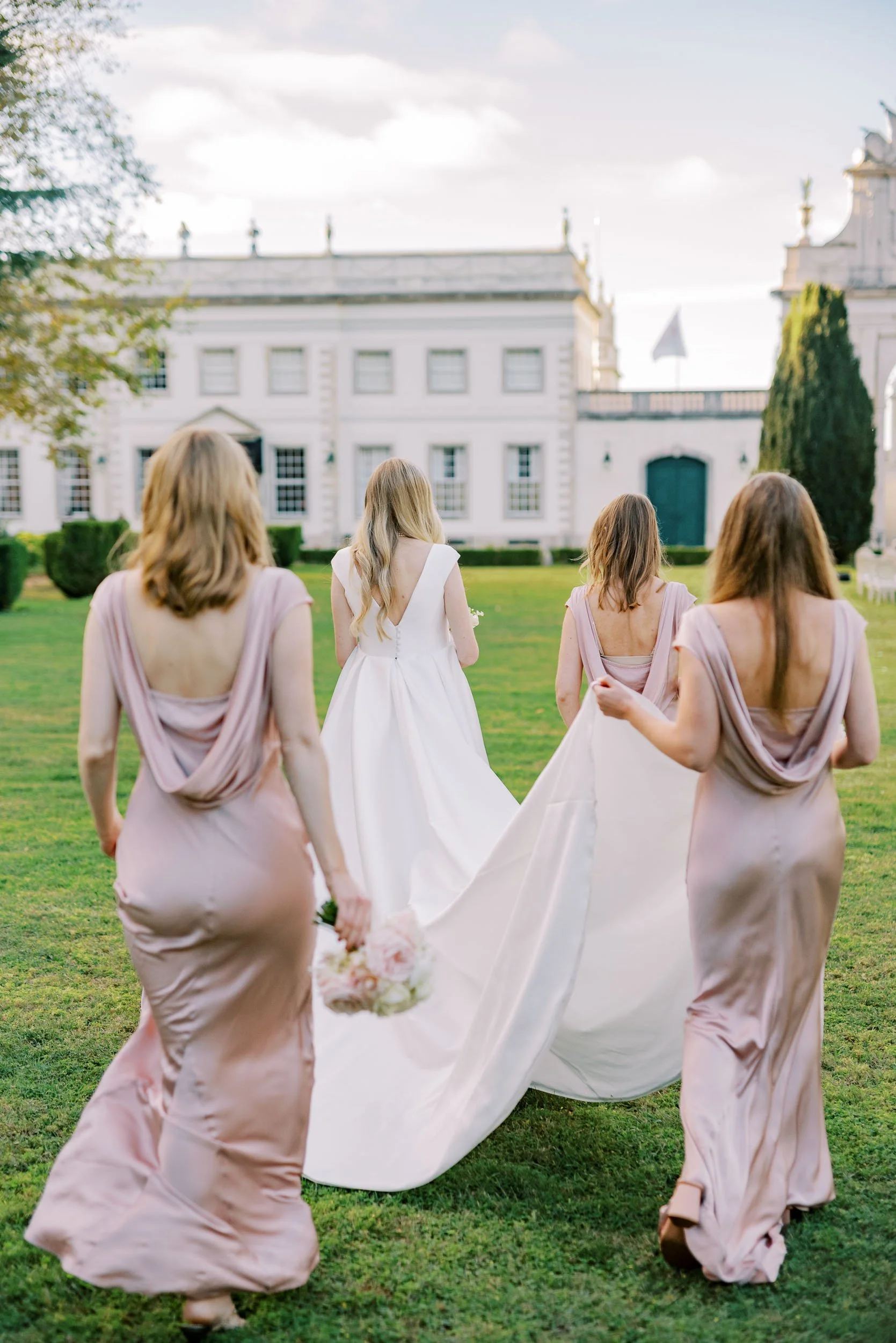 3Bride in a white wedding dress walking on a grassy lawn, accompanied by four women in matching light pink dresses, with Palácio de Seteais in the background