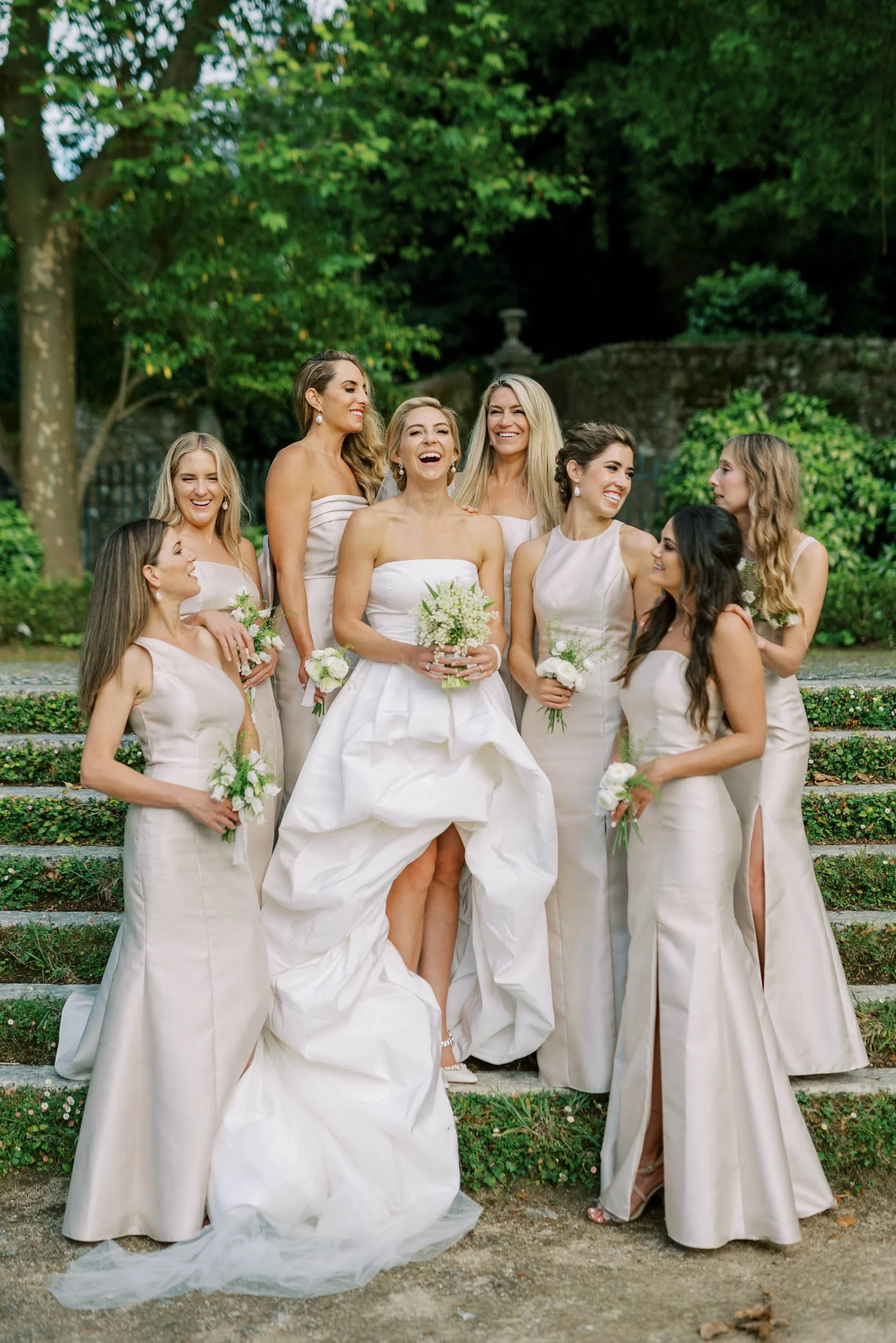 A bride and her bridesmaids stand outdoors on steps, all wearing cream-colored dresses, holding small white flower bouquets, with trees and greenery in the background at Palácio de Seteais in Sintra, Portugal.