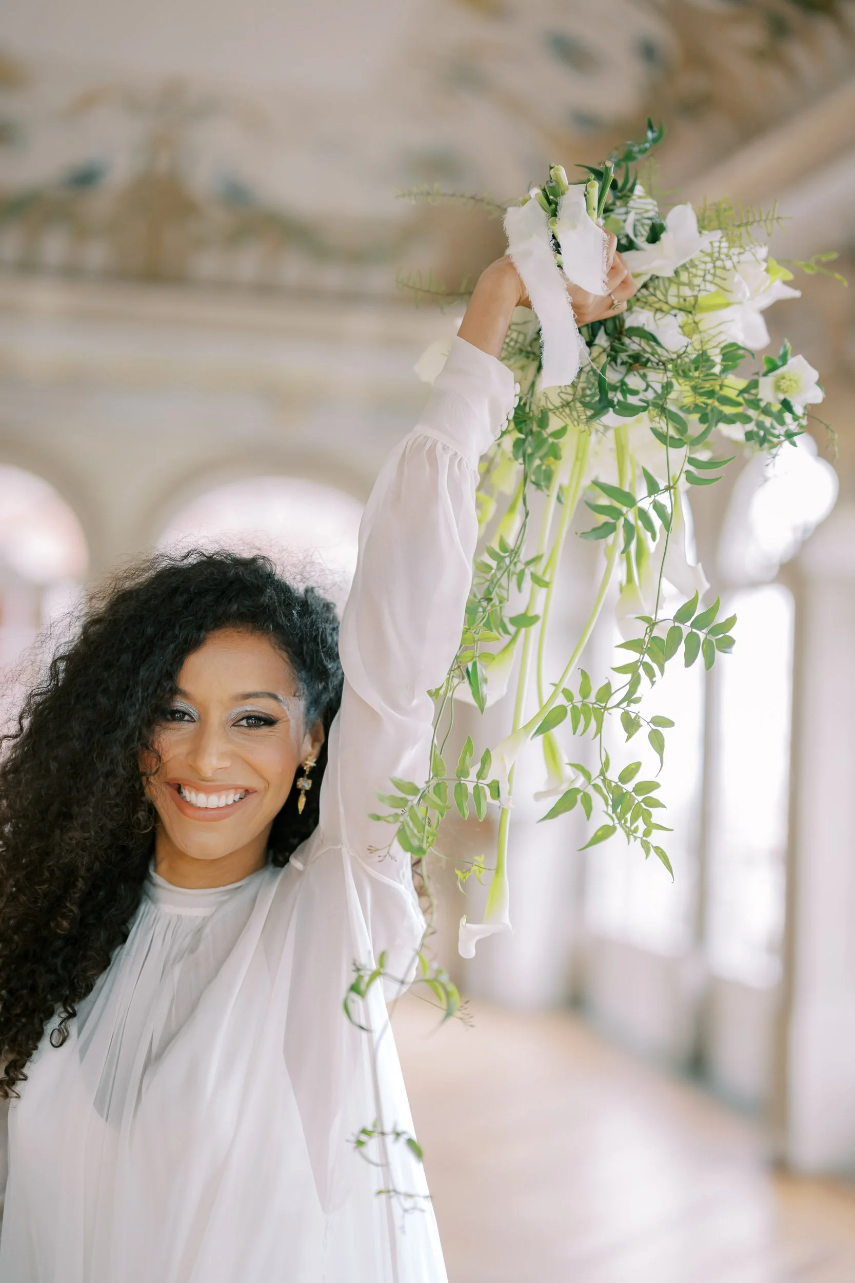 Bride with curly hair smiling and holding a bouquet of white flowers and greenery in a bright, elegant indoor setting.
