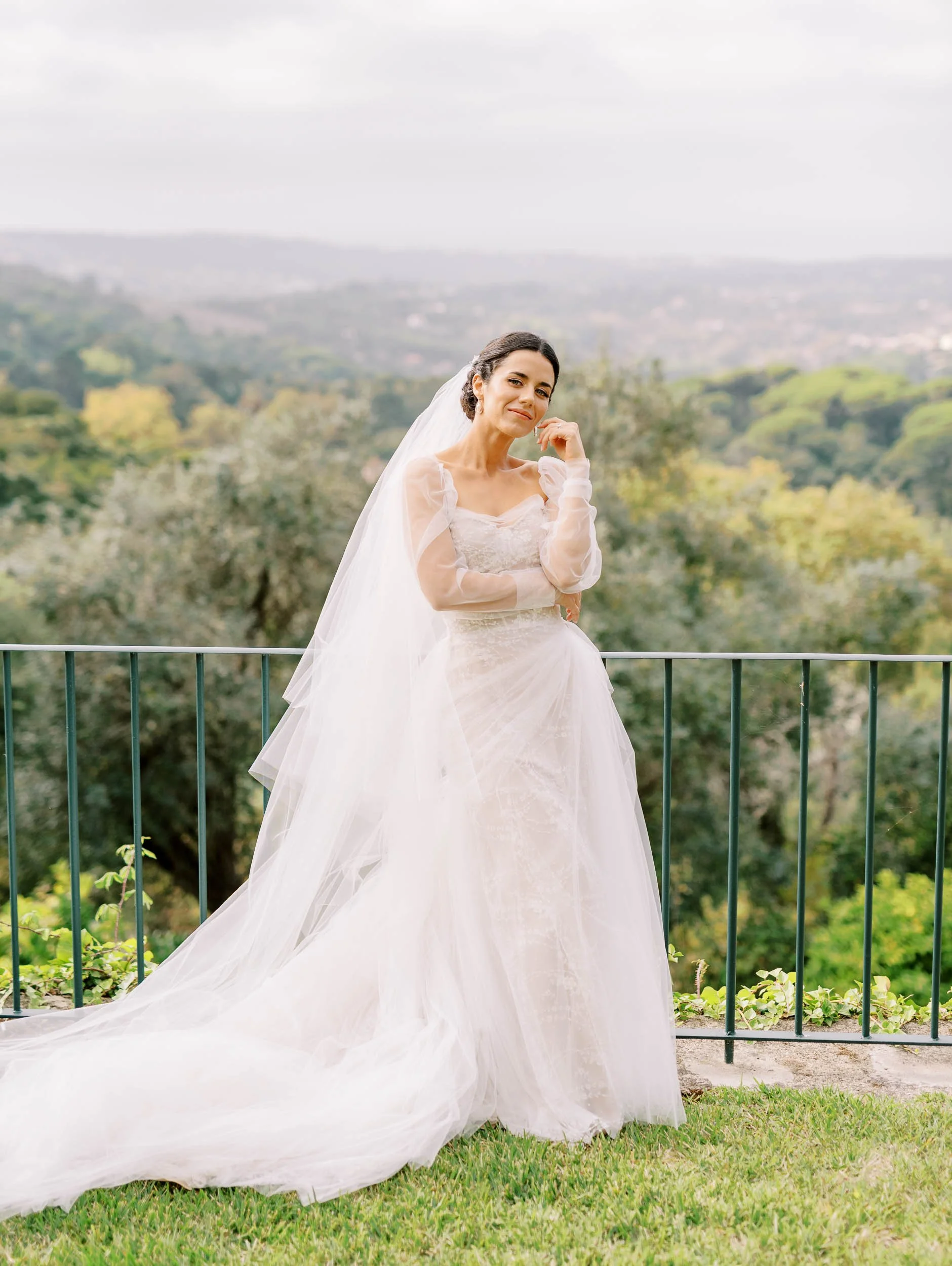 Bride in a white wedding gown with a long train and veil, standing outdoors on a grassy area with a scenic backdrop of trees and distant hills at Quinta da Bella Vista