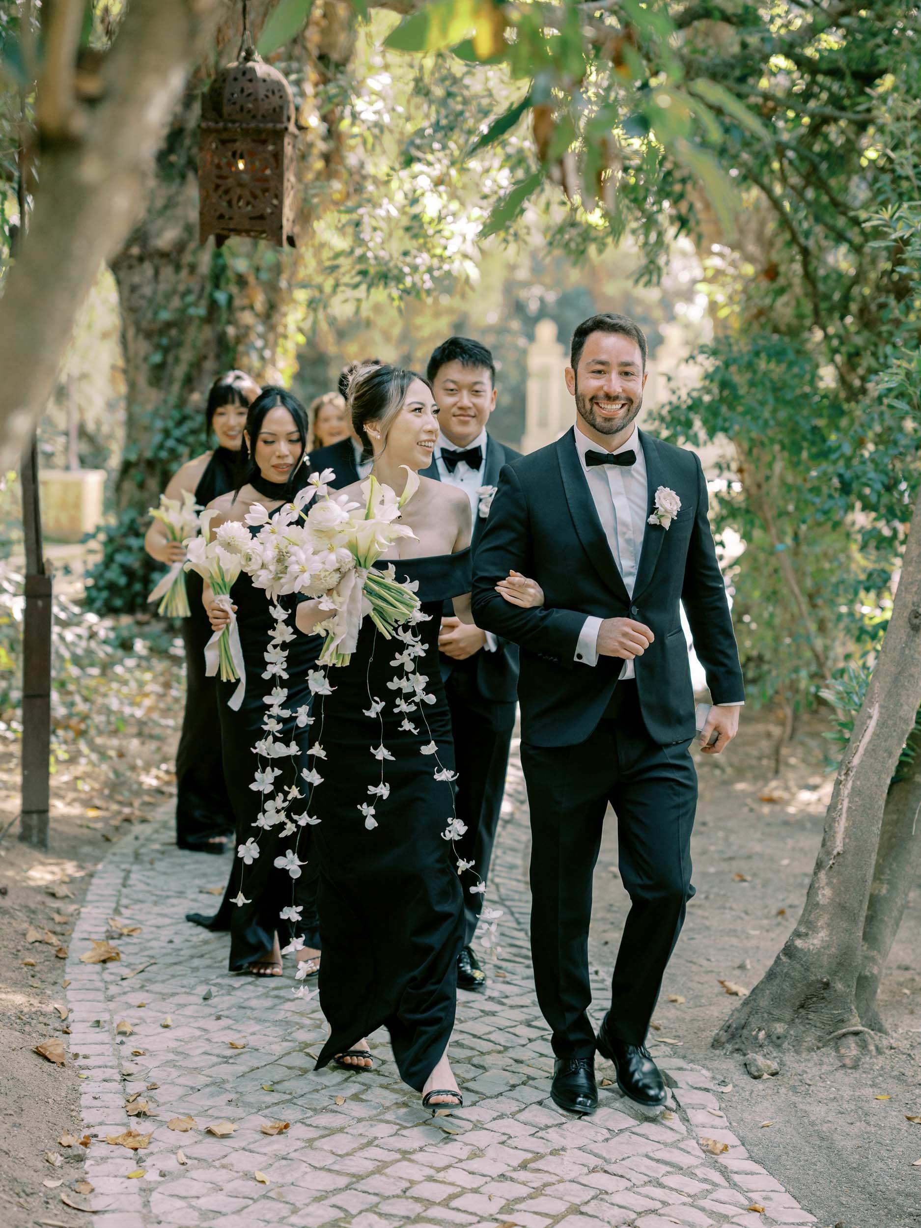 The wedding party dressed in formal attire walking on a cobblestone path in a garden, with a man in a tuxedo leading, holding hands with a woman carrying a bouquet of flowers, surrounded by lush trees at Quinta da Bella Vista.