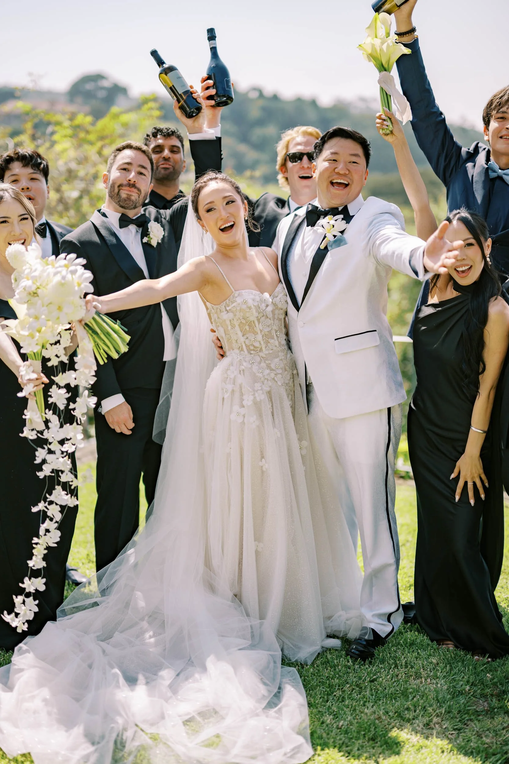 Group of wedding guests celebrating outdoors, with the bride and groom at the center, holding wine bottles and flowers, all smiling and joyful.