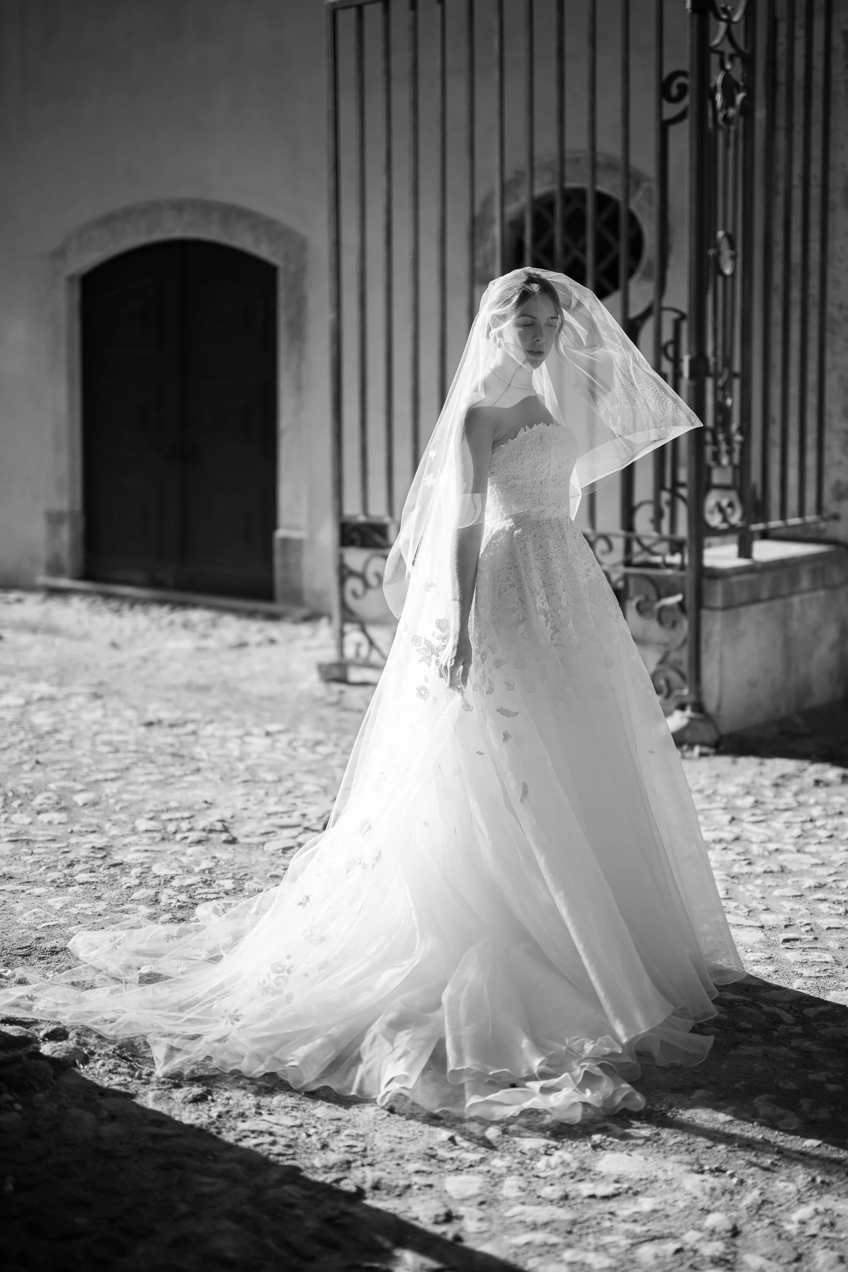 A bride in a strapless wedding gown and veil standing outdoors on a stone-paved path, with a building and gate in the background, captured in black and white at Palácio do Correio-Mor
