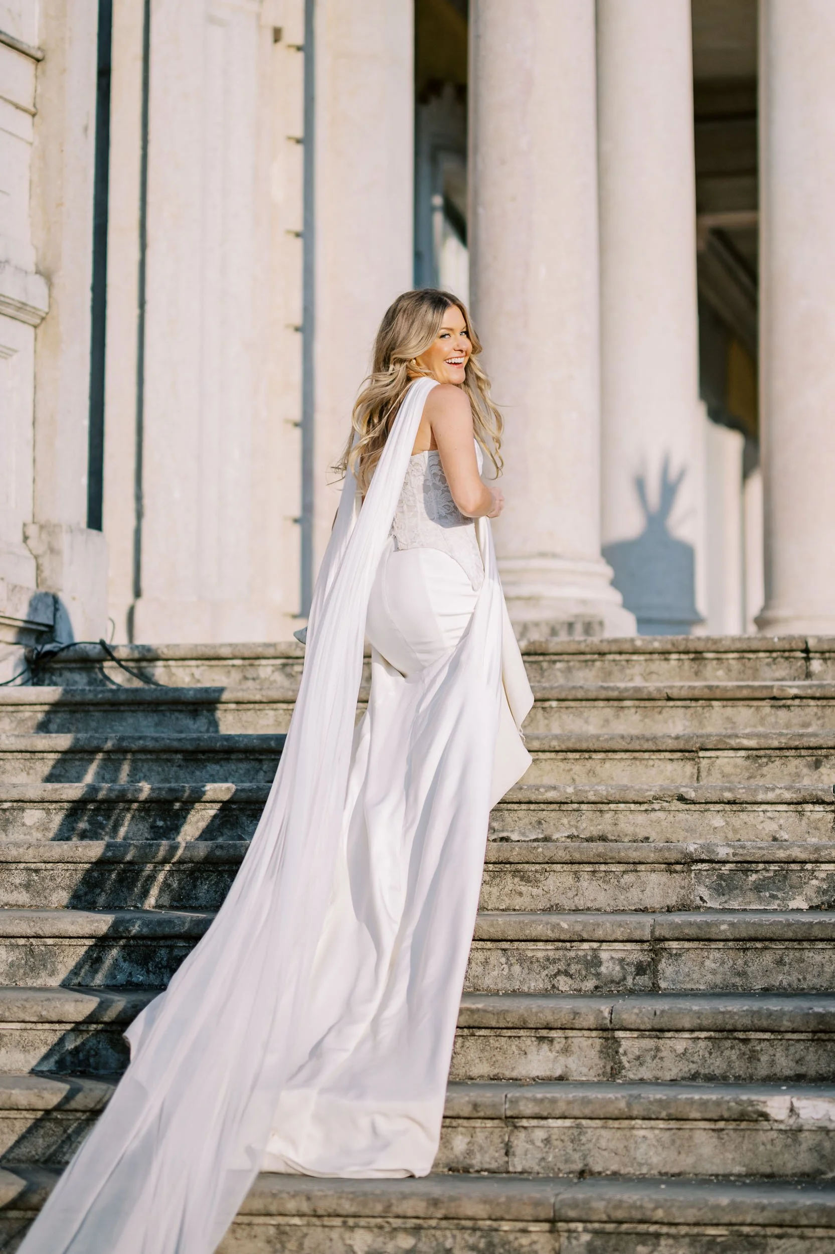 A woman in a white wedding dress with a long train stands on stone steps outside a building with large columns, smiling and looking back at Palácio de Queluz in Lisbon