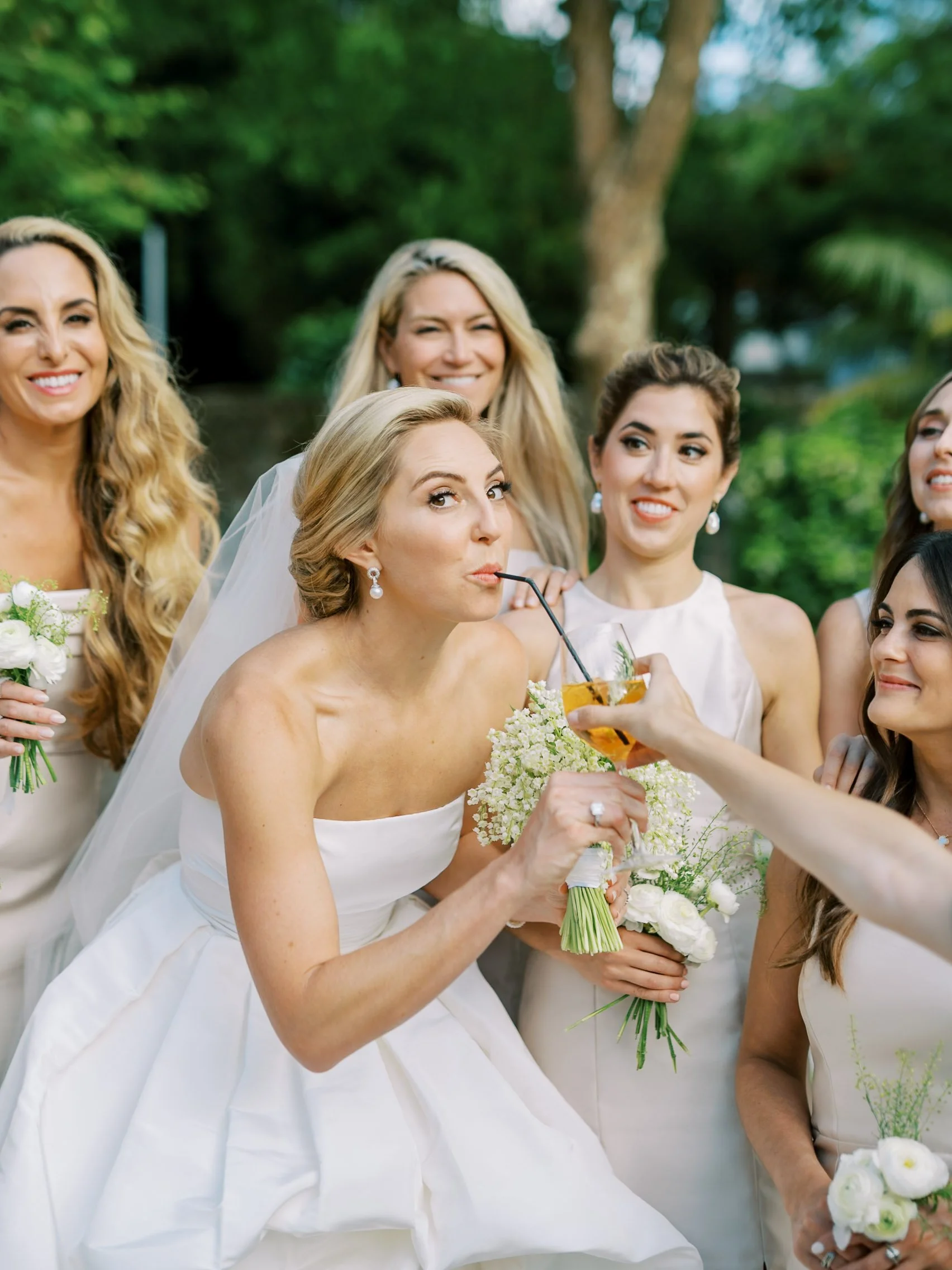 A bride in a white wedding dress is sipping a drink through a straw held by another person at a wedding reception with bridesmaids and greenery in the background.
