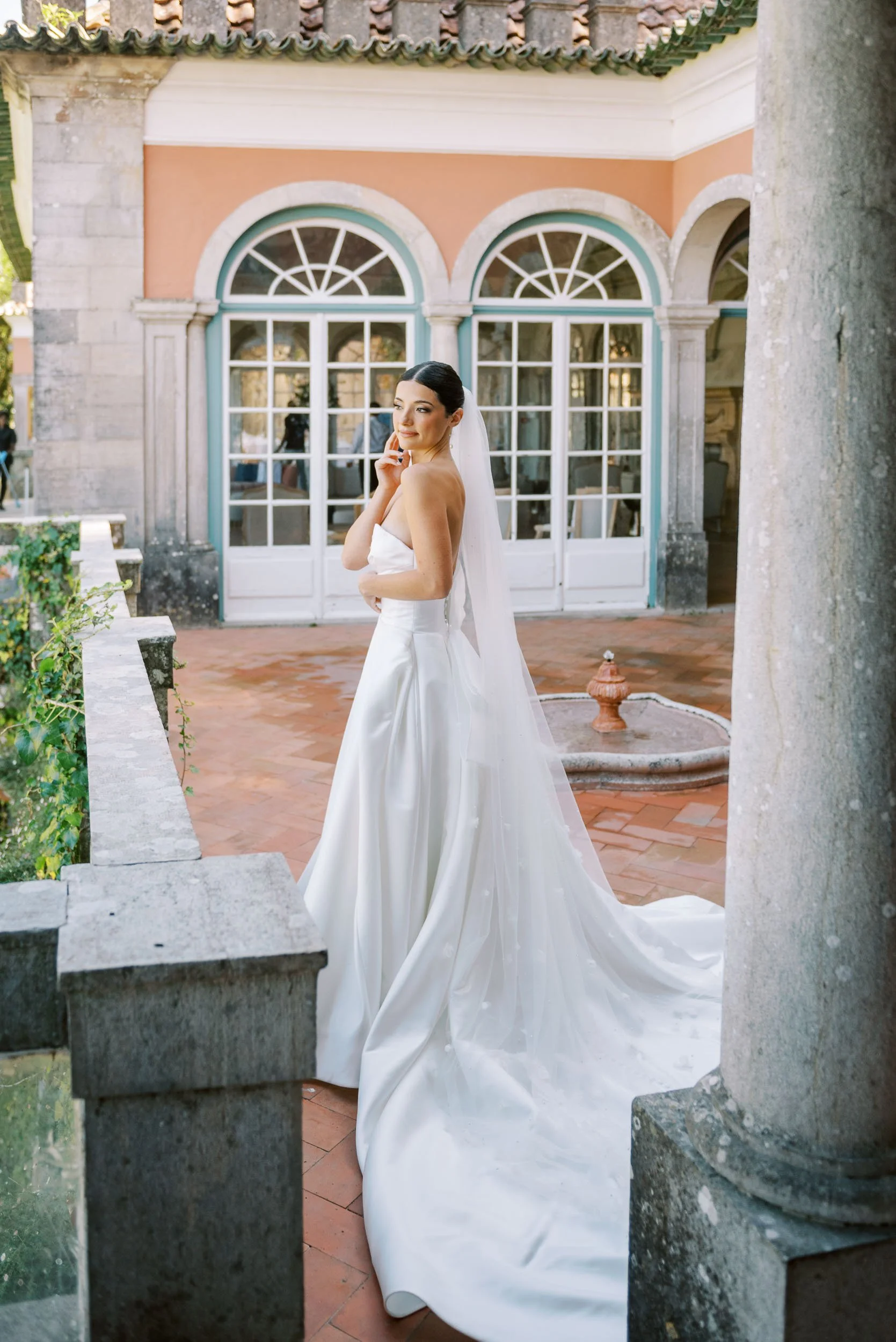 A bride in a white wedding gown with a long train and veil, standing outdoors near a stone fountain and a building with large arched windows and pastel-colored walls at Casa dos Penedos in Sintra, Portugal.