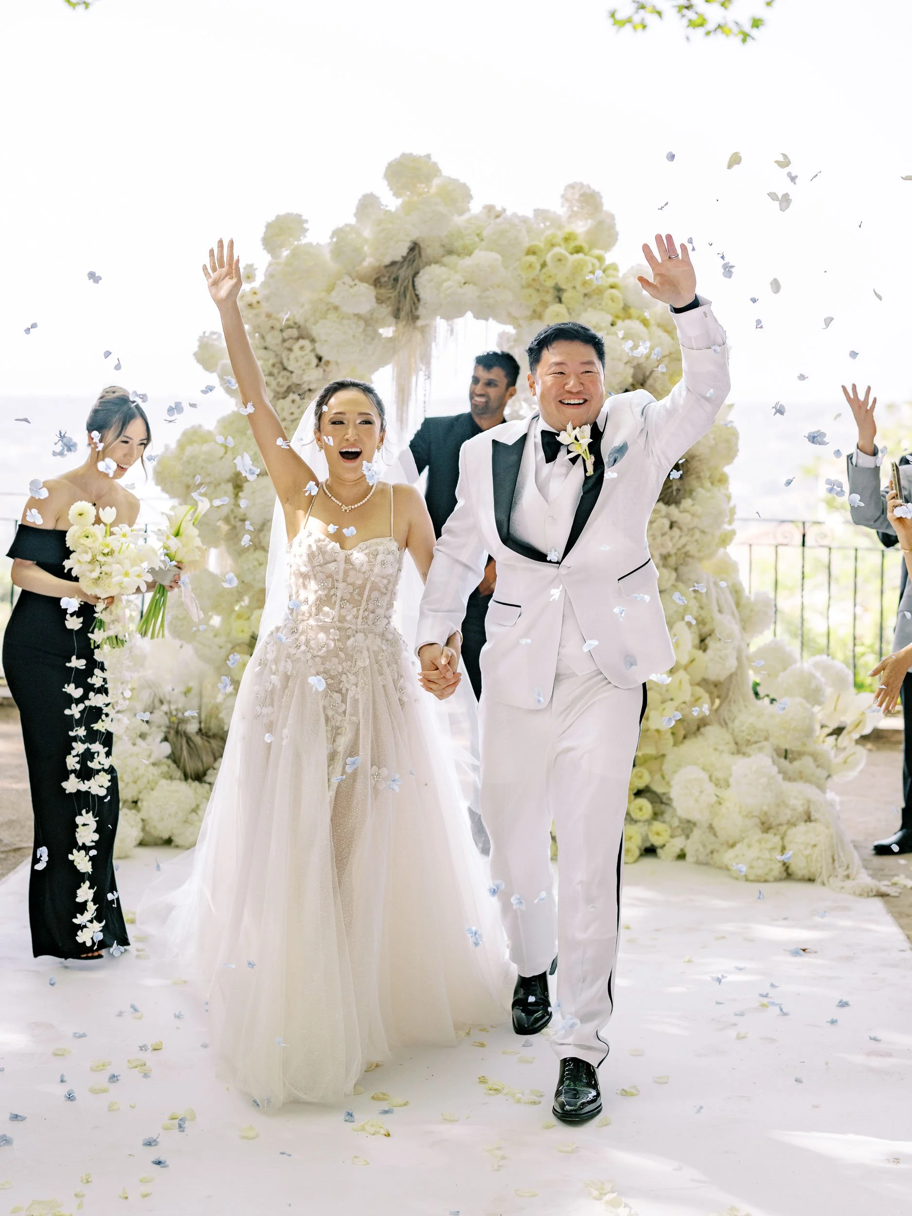 A wedding celebration with a bride and groom holding hands and smiling, surrounded by flower decorations and guests, as confetti falls at Quinta da Bella Vista