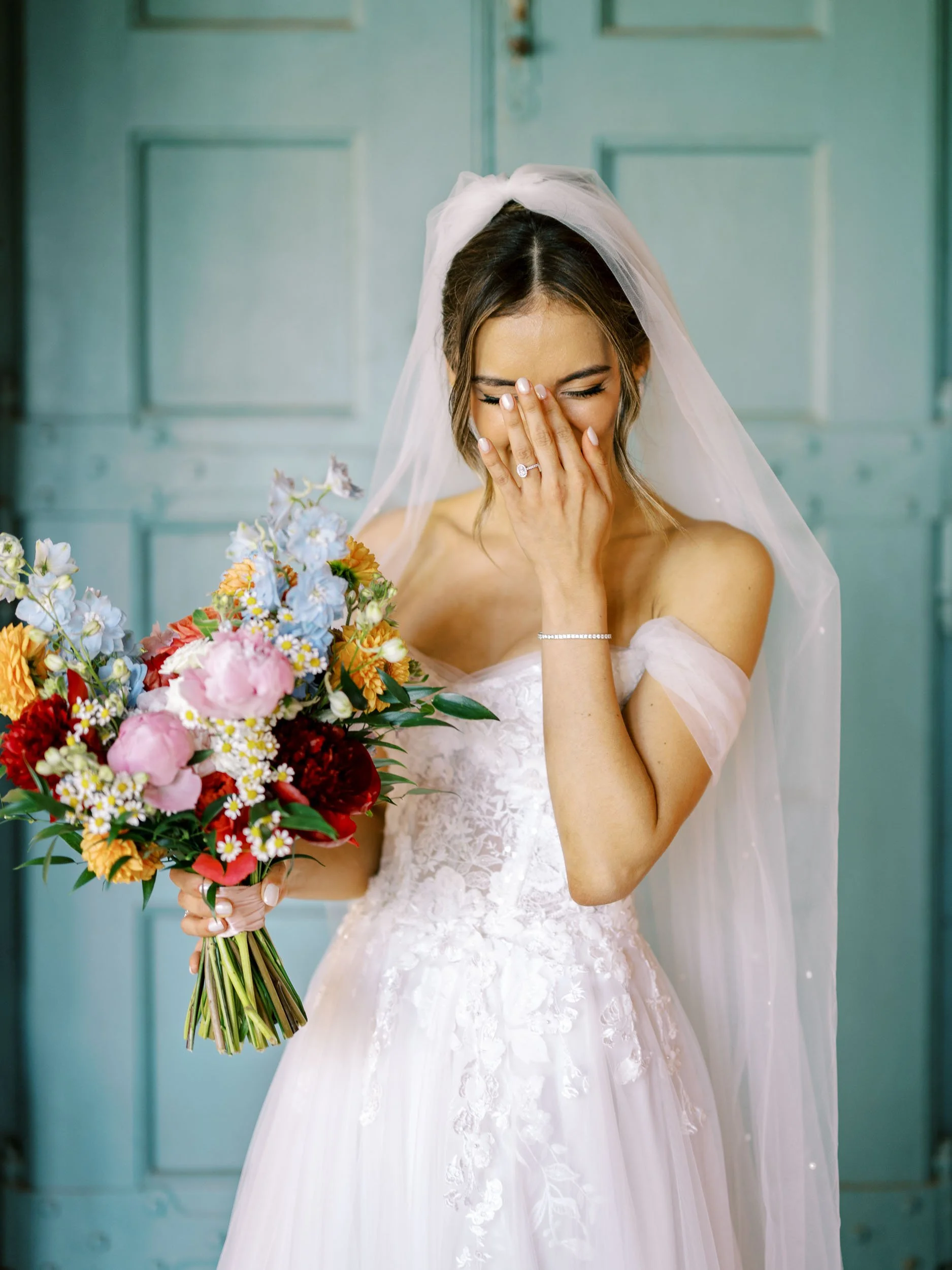 A bride in a white wedding dress holding a colorful bouquet of flowers, covering her face with her hand and smiling, standing in front of a teal door.