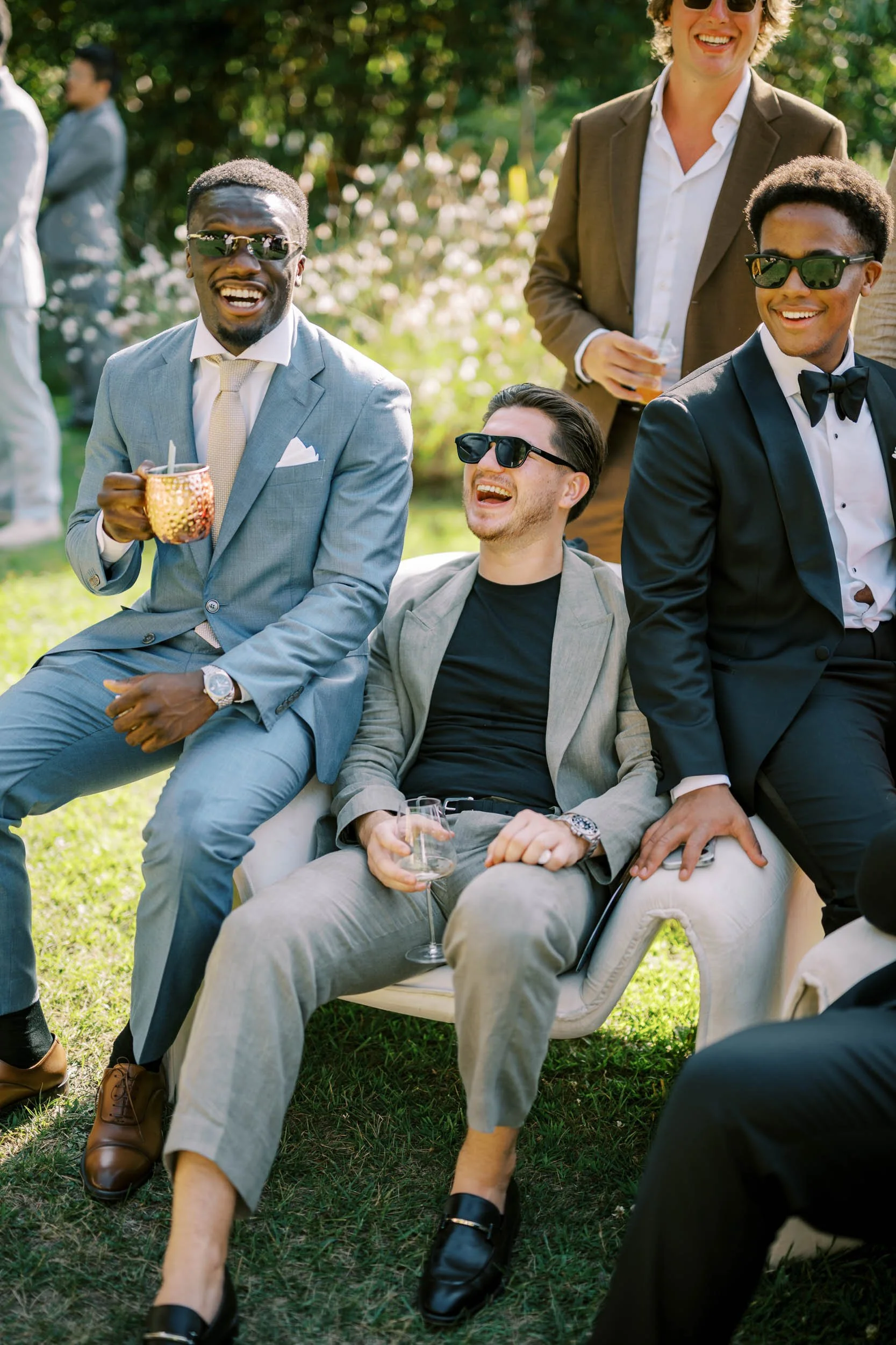 Group of wedding guests dressed in formal attire, enjoying drinks and smiling at an outdoor event on a sunny day at Quinta da Bella Vista in Sintra.