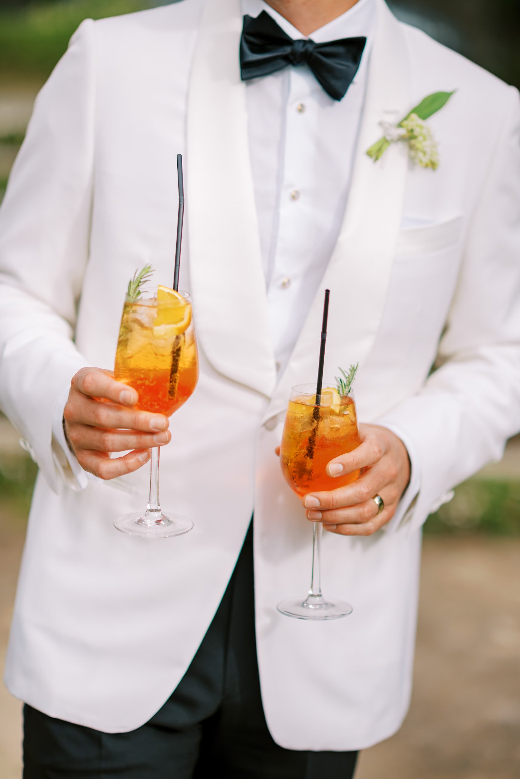 Groom in white tuxedo holding two Aperol spritz cocktails with orange slices and garnishes.