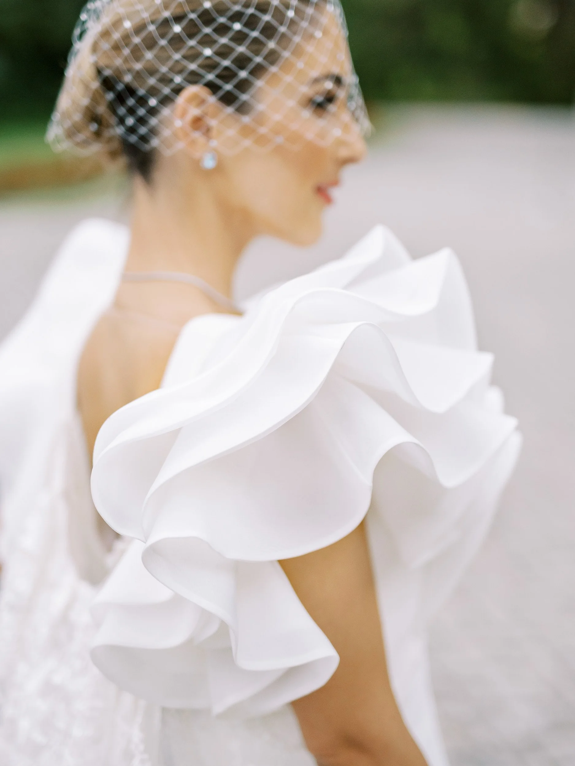 A woman wearing a white dress with large ruffled sleeve, a birdcage veil, and pearl earrings.