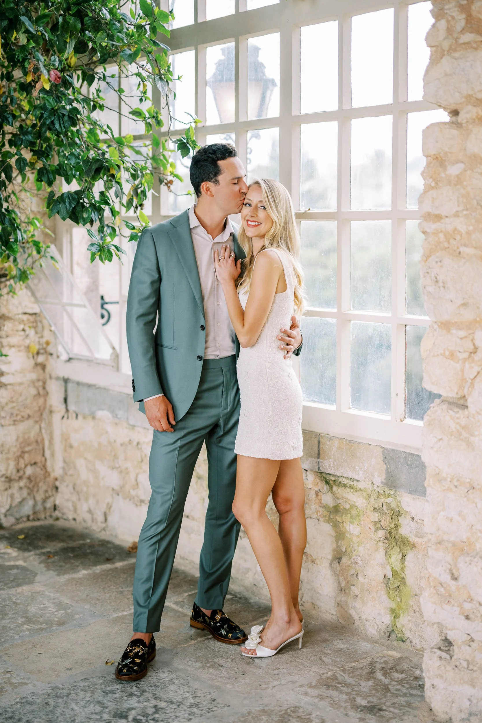 A smiling wbride in a white dress and white heels standing close to the groom in a suit, with the man kissing her on the forehead, inside a bright, rustic space with large windows and stone walls at Forte da Cruz in Cascais, Portugal.