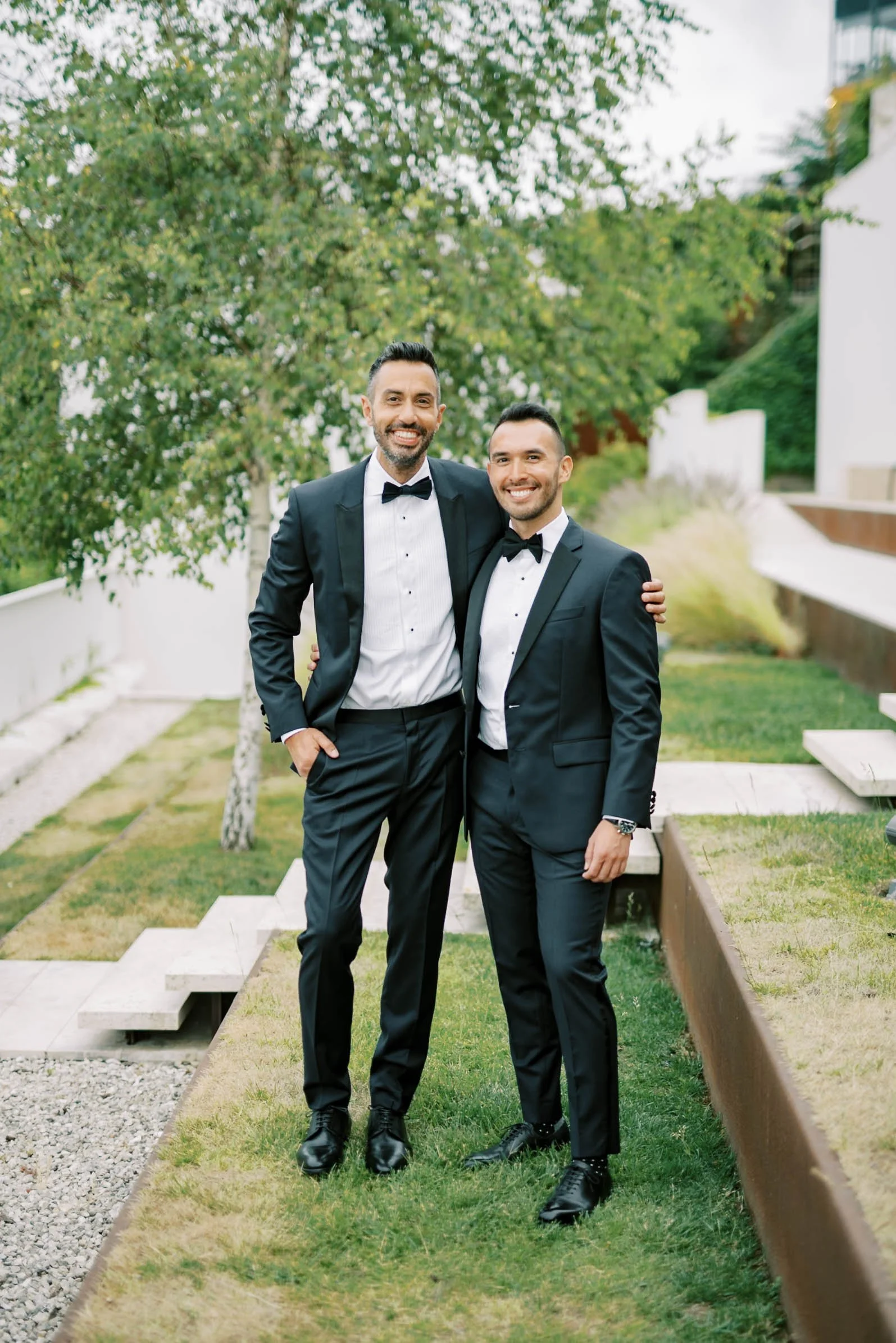 Two grooms in black tuxedos with white shirts and black bow ties standing outdoors on a grassy area, smiling at the camera, with trees and a modern white building in the background.