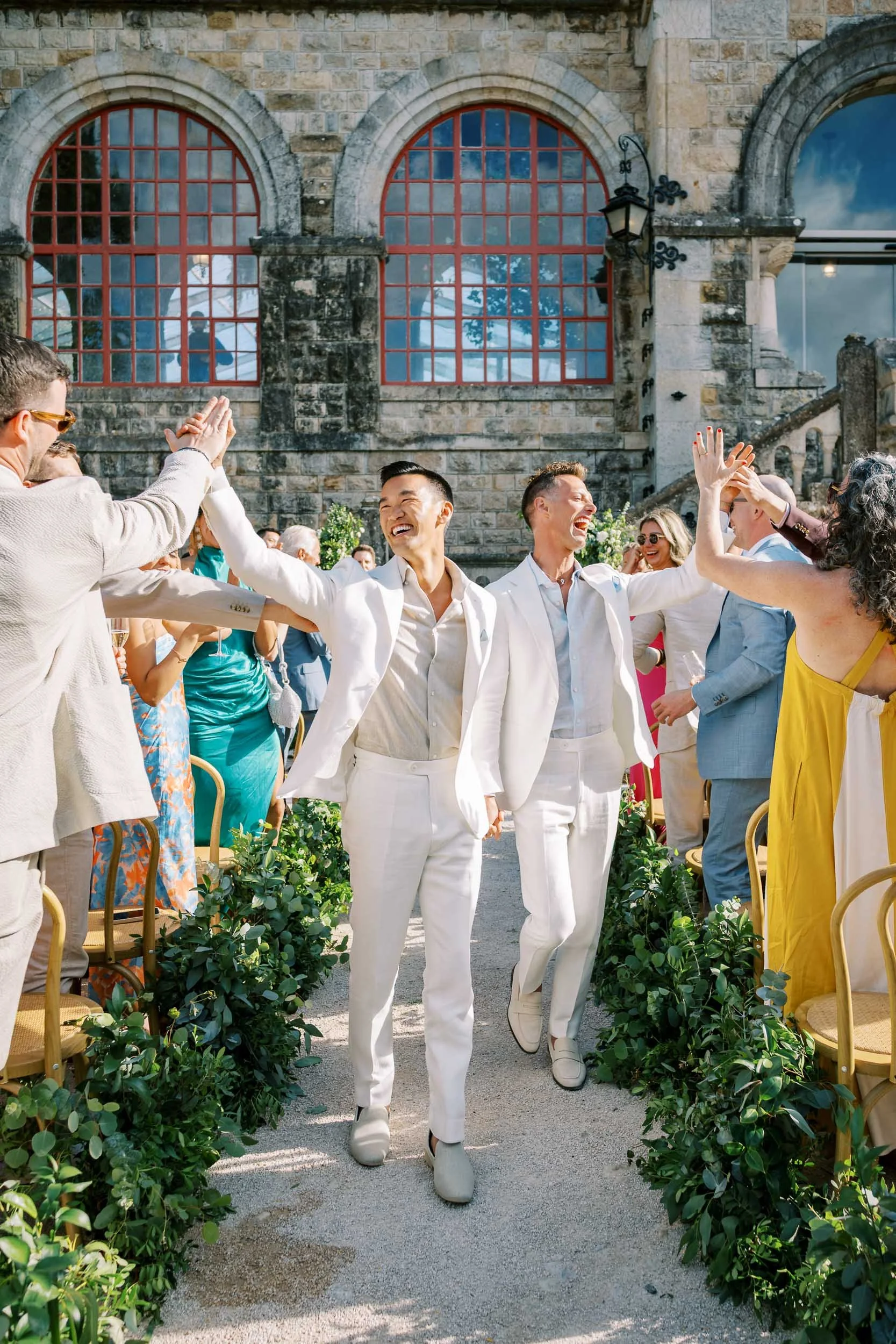 Celebrating wedding at outdoor venue with two grooms in white suits holding hands and high-fiving, surrounded by guests in colorful attire.