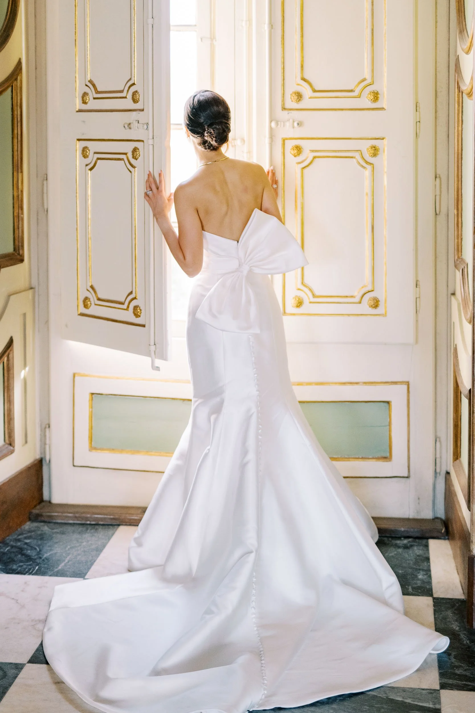 Bride in a strapless white wedding gown with a large bow at the back, standing by open shutters, gazing outside in a room with gold-accented white walls at Palácio de Queluz in Lisbon