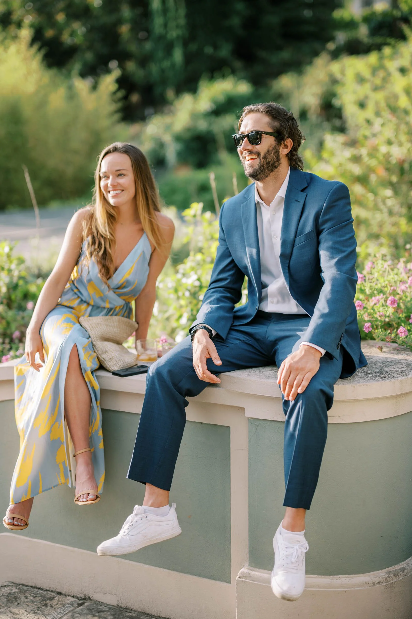 A man in a blue suit and sunglasses sits on a ledge, smiling, next to a woman in a light blue and yellow dress, also smiling, as they enjoy a sunny outdoor setting with greenery and pink flowers.