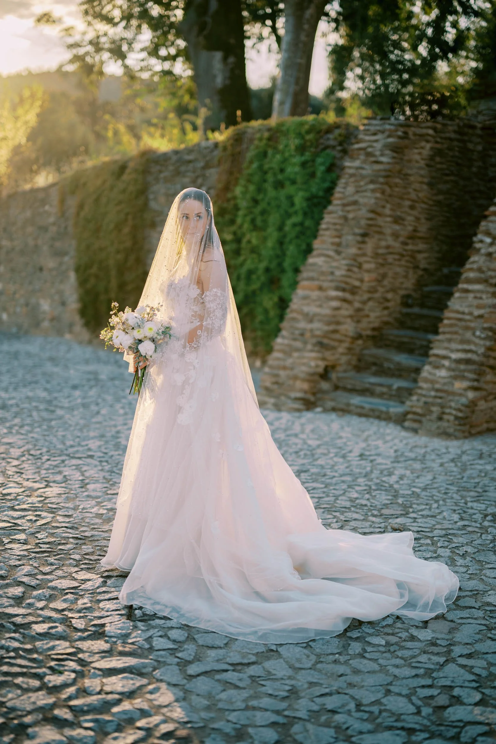 A bride in a white wedding dress and veil holding a bouquet of white flowers standing on a cobblestone path near an old stone staircase and a grassy wall, with trees and sunlight in the background.
