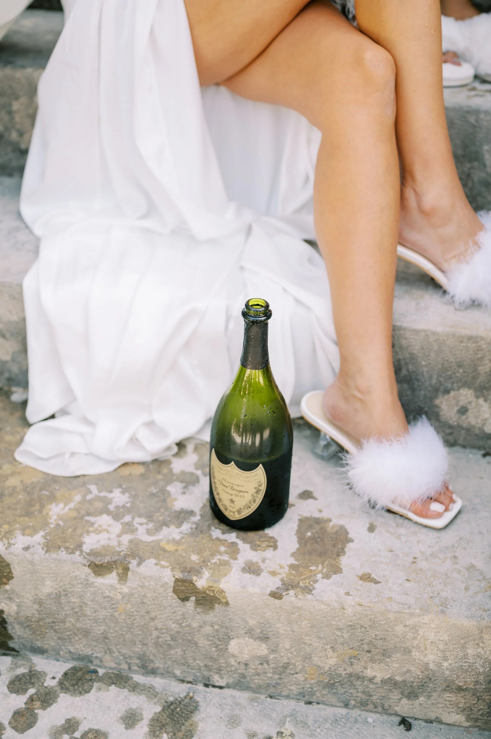 A bride wearing a white dress and fluffy white slippers is sitting on concrete steps with a partially empty champagne bottle on the ground nearby.
