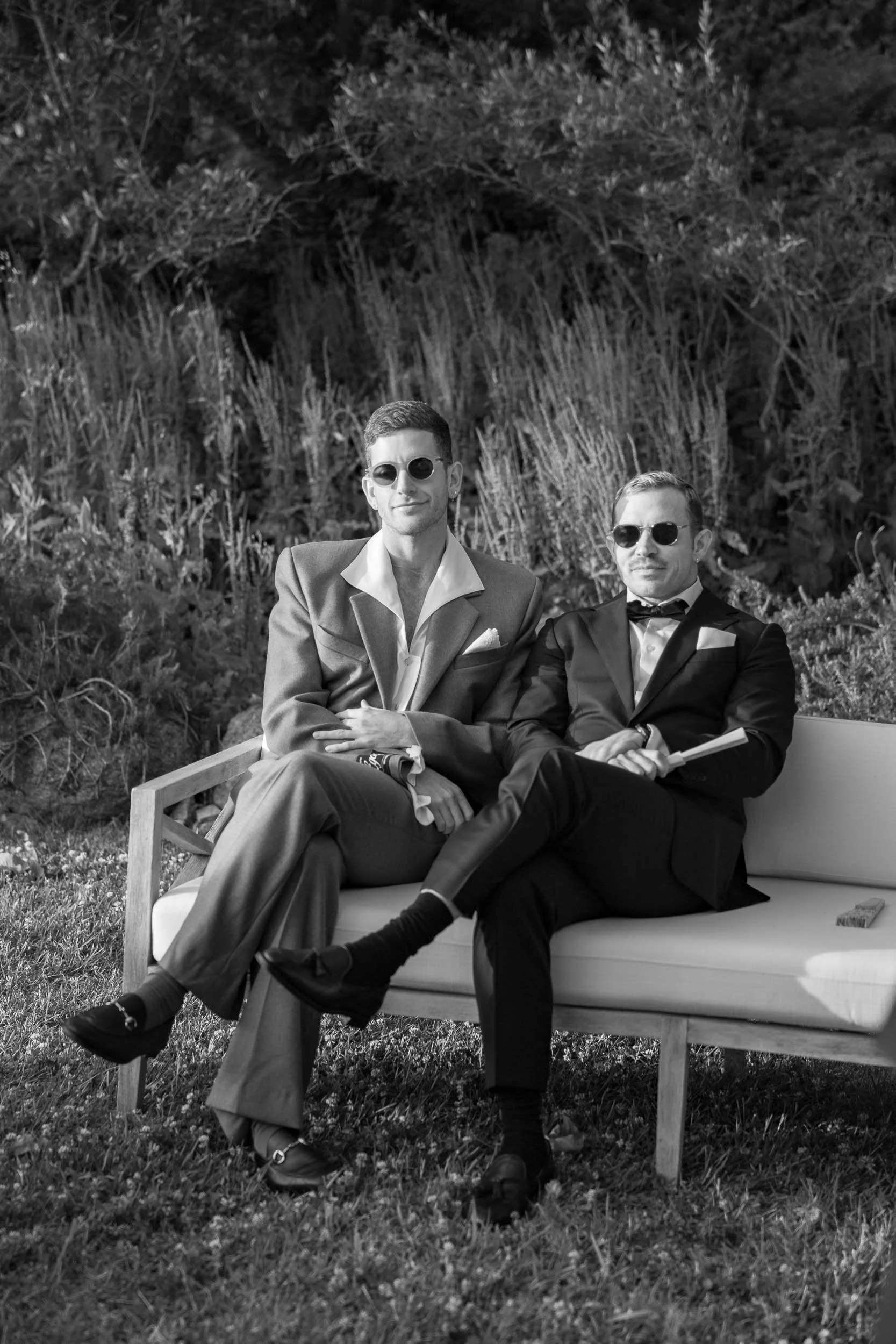 Two wedding guests in suits sitting on a bench outdoors, wearing sunglasses, with tall grass and bushes in the background.