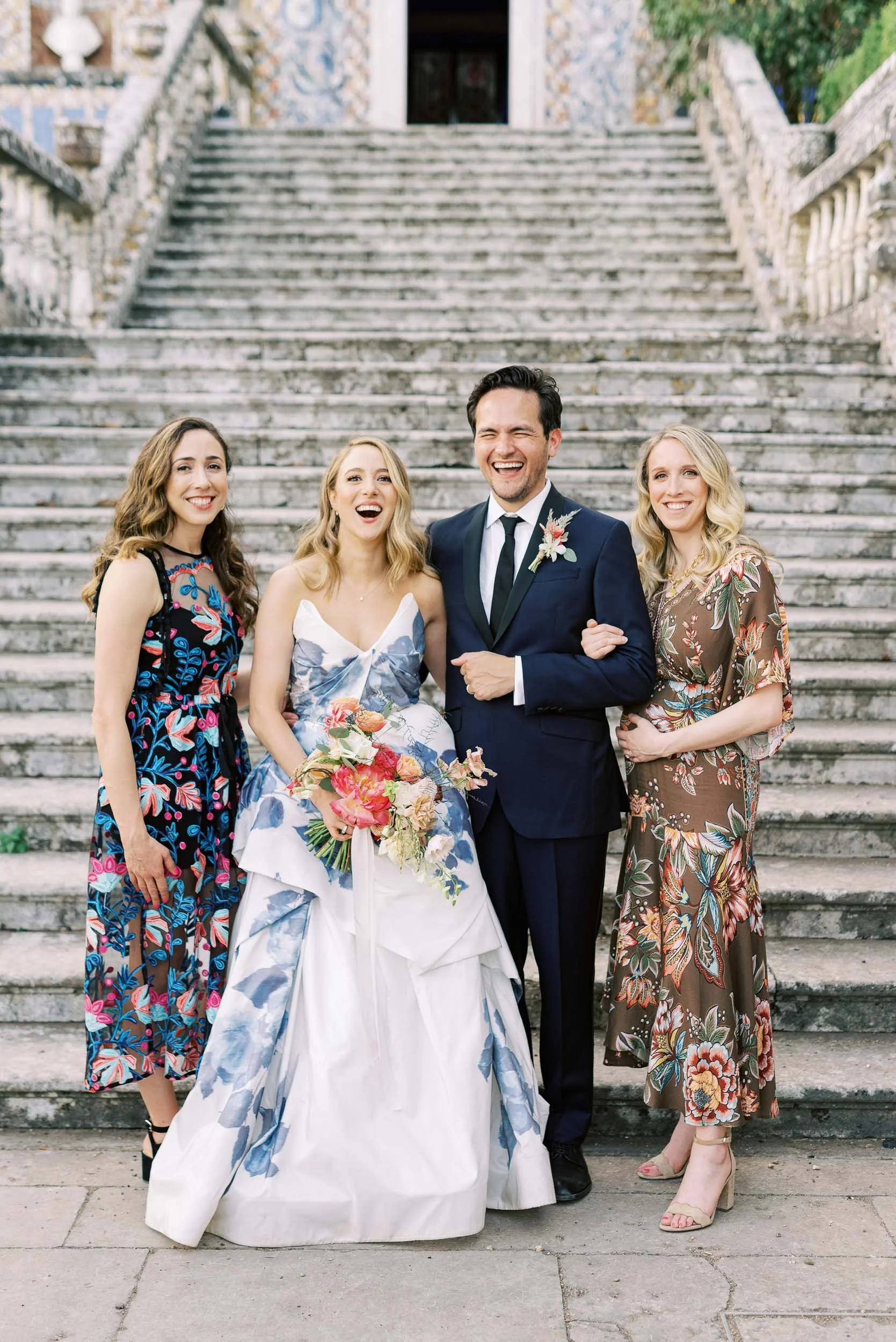 Group of four smiling people standing on stone steps outdoors, with two women in floral dresses and a man in a dark suit and tie, one woman holding a bouquet of flowers in front of her, during a wedding celebration.