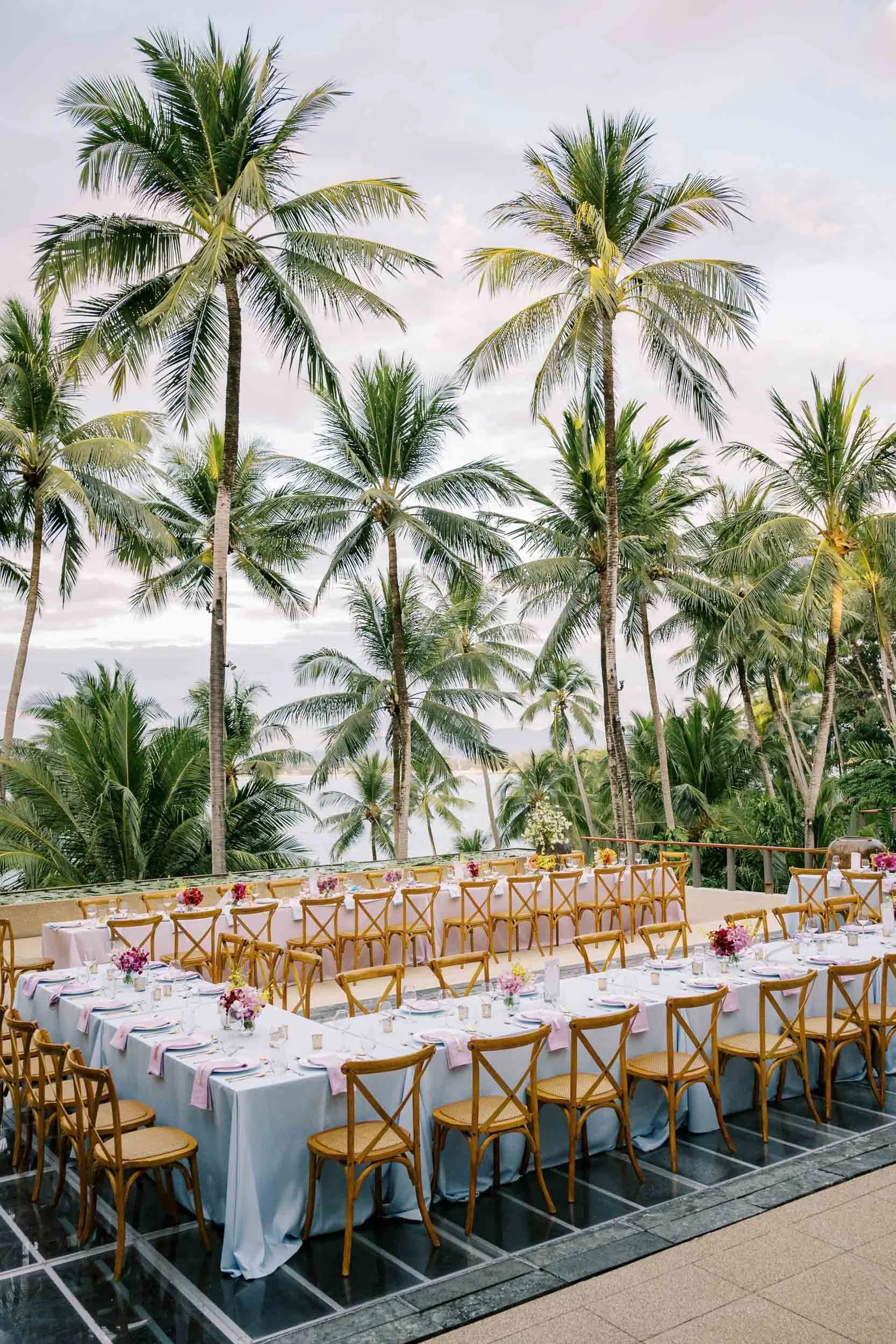 Dinner table set outdoors at Amanpuri hotel in Phuket surrounded by palm trees, with a view of the ocean in the background, decorated with flowers and pink table napkins.