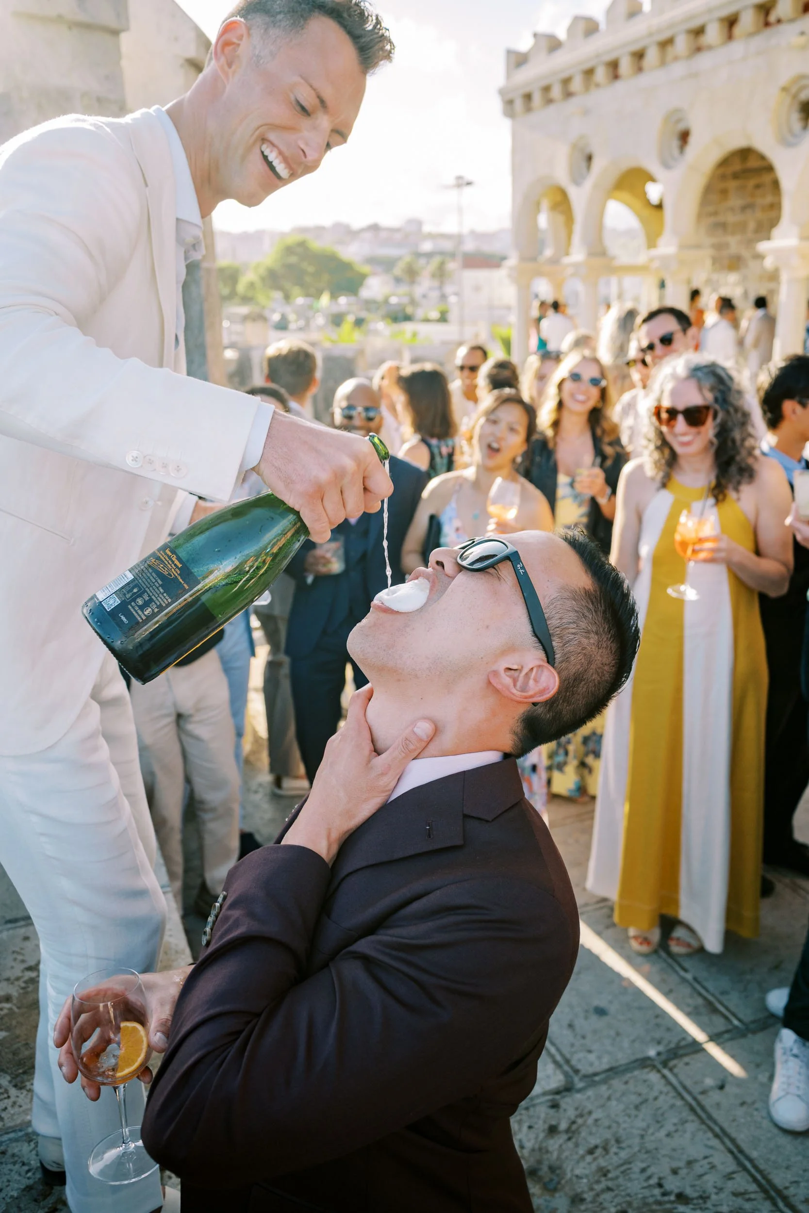 A groom in white suit pours champagne into a guest's mouth during a lively outdoor celebration with a crowd of smiling people.