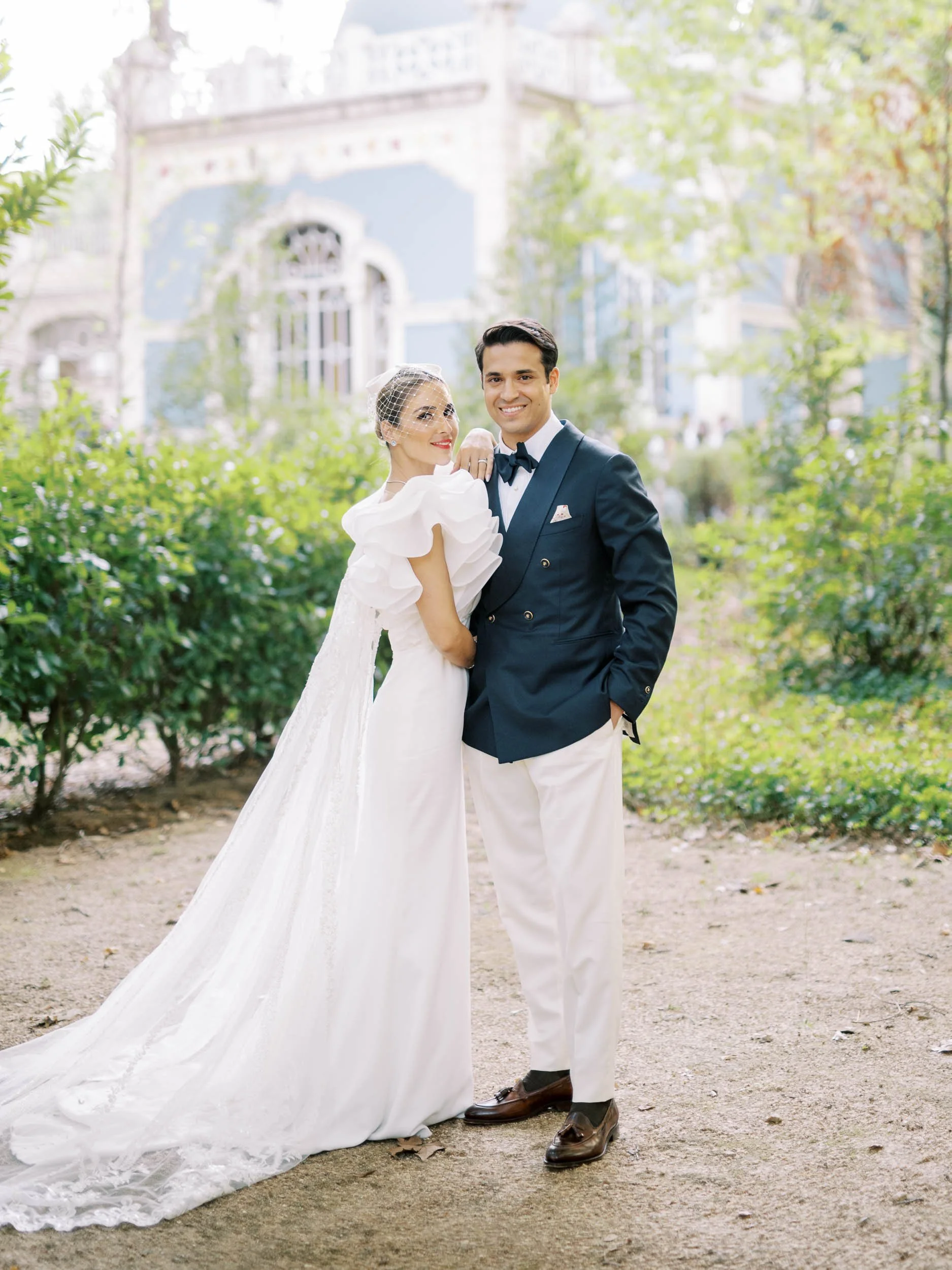 A bride in a white wedding gown with ruffled sleeves and a birdcage veil, and a groom in a navy blue tuxedo with a bow tie, posing together in a garden with greenery and a historic building in the background.