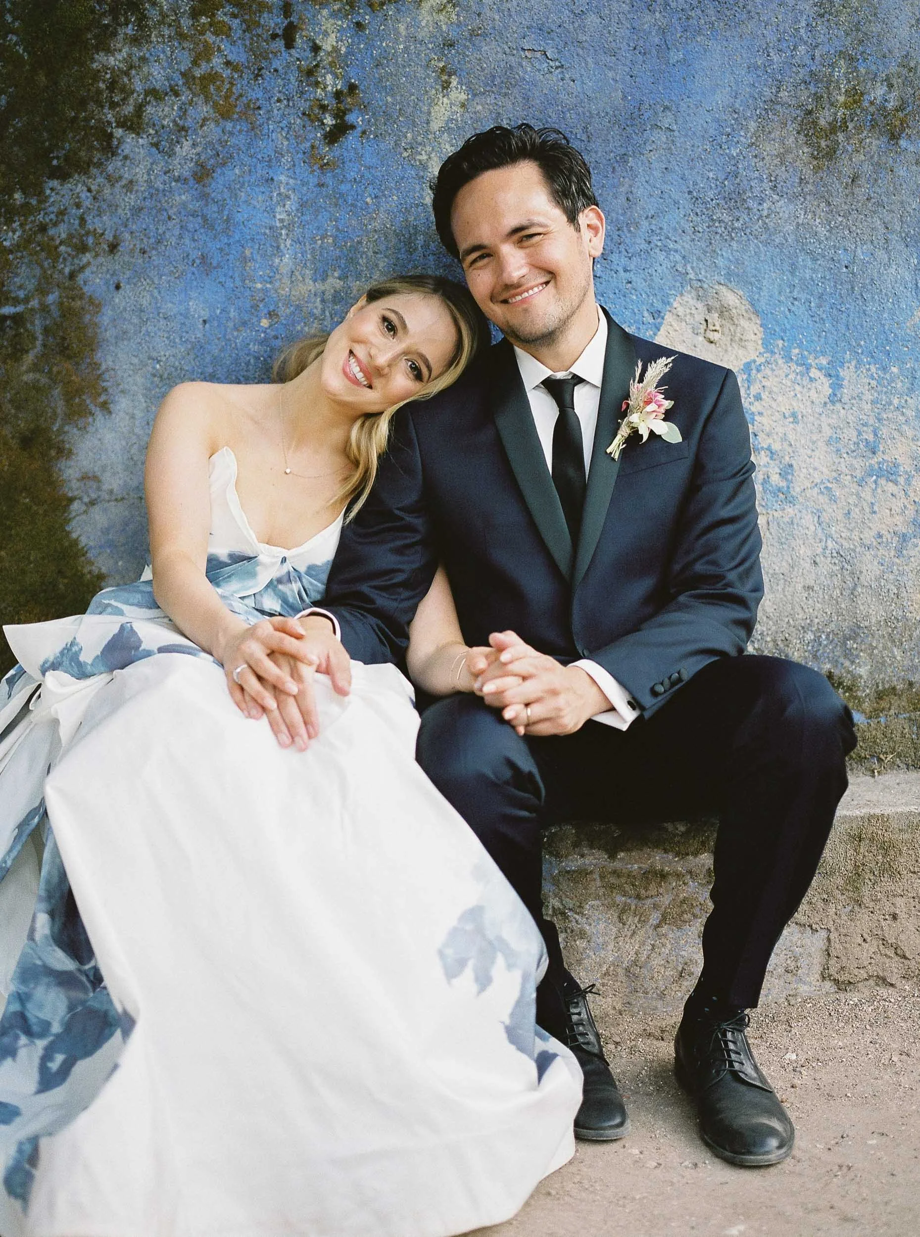 A smiling bride and groom sitting close together outdoors against a textured blue wall. The bride leans her head on the groom's shoulder, her hand resting on his knee, wearing a strapless wedding gown. The groom is dressed in a navy blue suit with a 