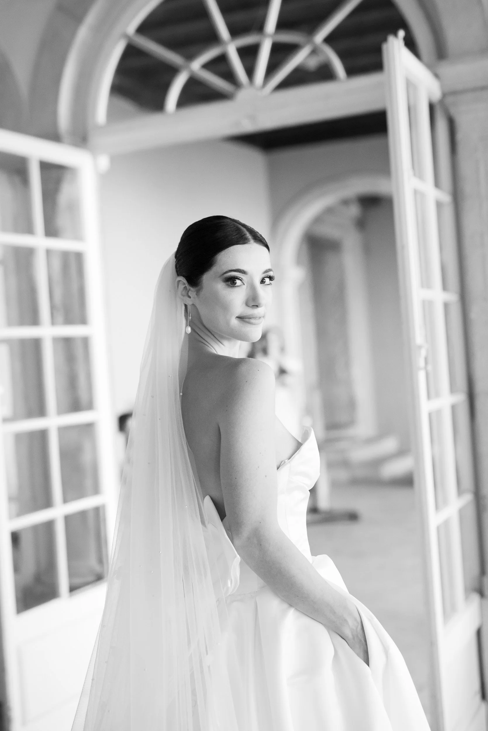 Black and white photo of a bride in a wedding gown with a veil, standing near open double doors with glass panes and arches in a historic building.