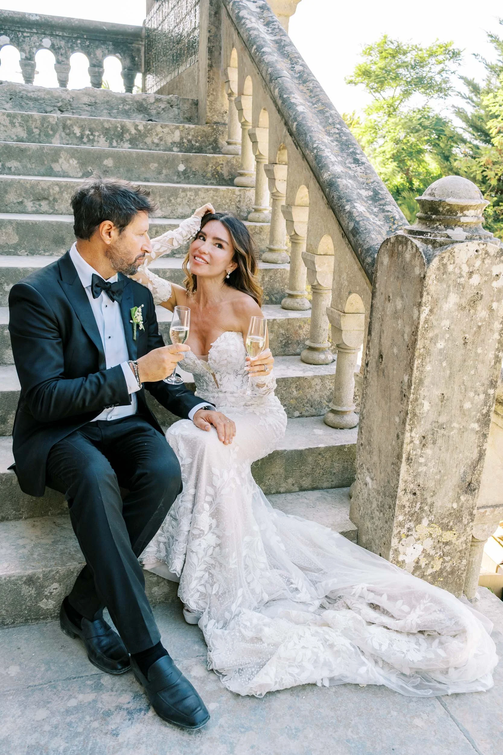 A couple dressed in wedding attire sitting on stone steps, toasting with champagne glasses, with a stone staircase and greenery in the background.