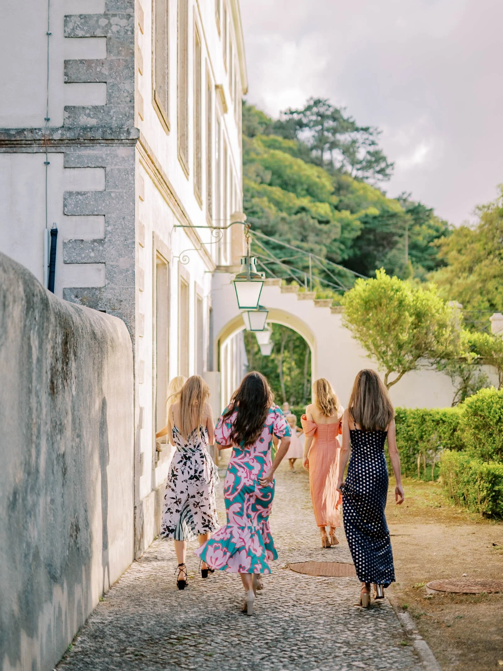 Group of bridesmaids walking along a paved path next to a white building with lamps overhead, surrounded by greenery and trees, in a scenic outdoor setting at Palácio de Seteais in Sintra