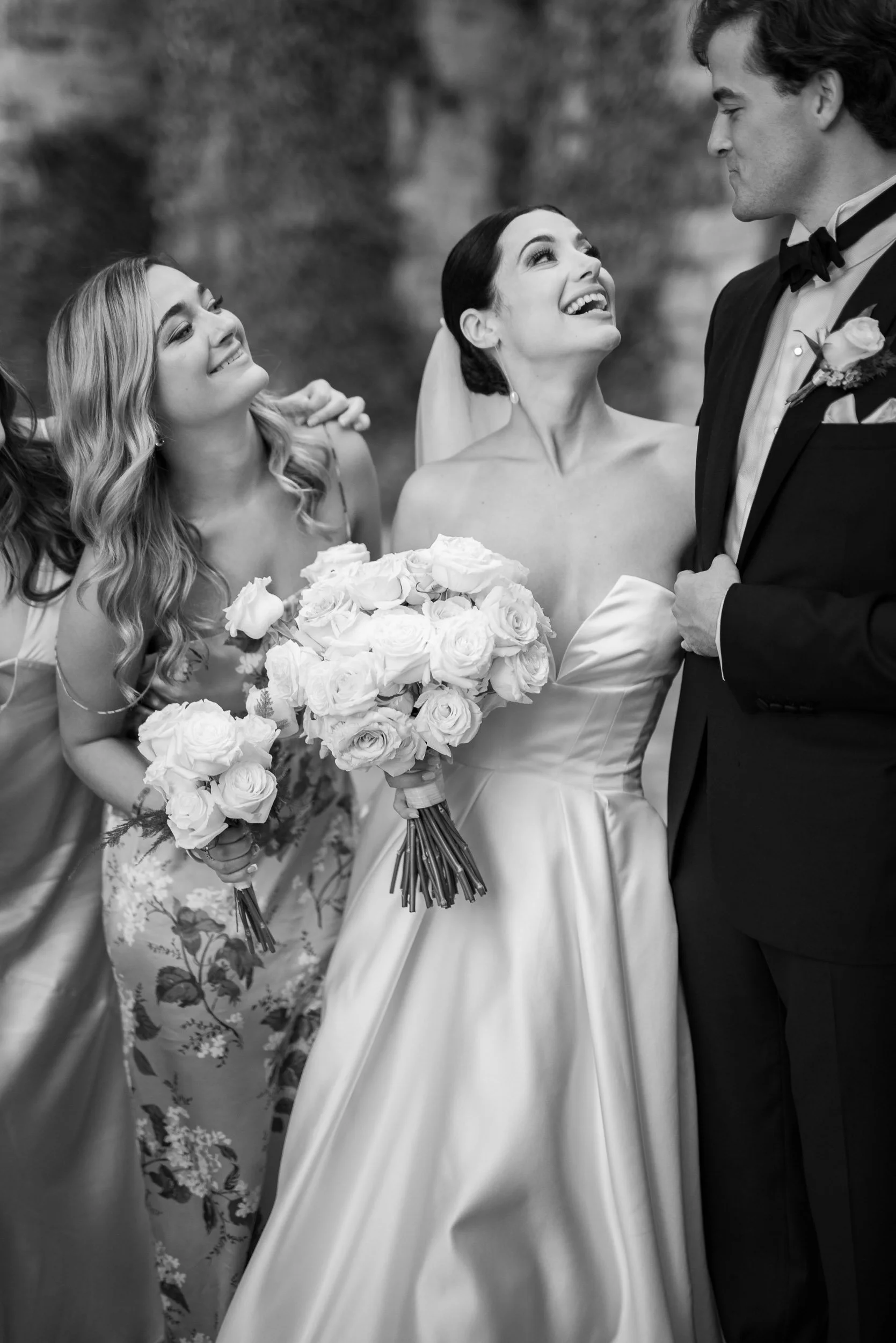 A black and white photo of a bride in a wedding gown looking up happily at a groom in a tuxedo, holding a bouquet of roses, while a bridesmaid next to her smiles and observes.
