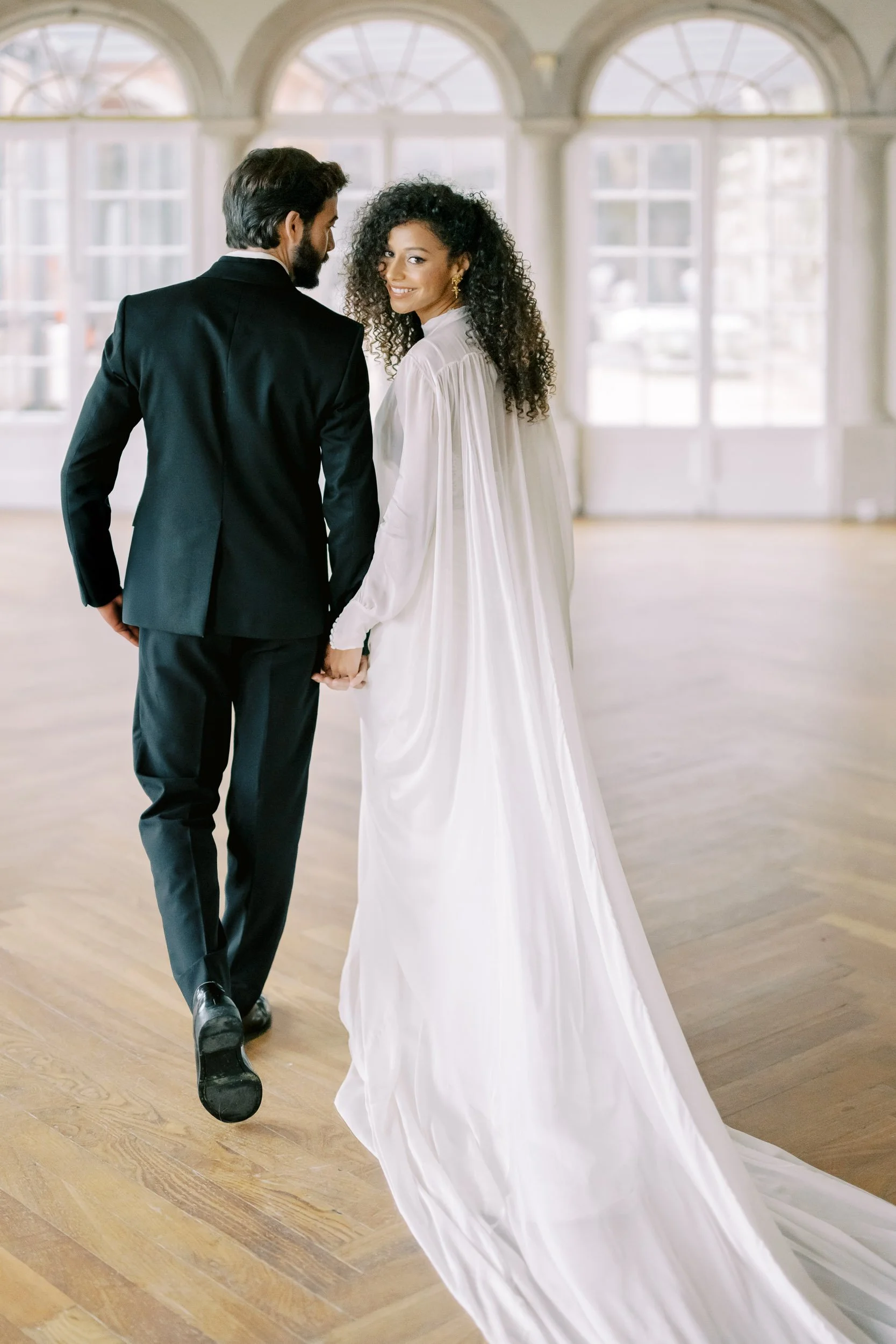 A couple dressed in wedding attire holding hands, with the bride in a white gown and the groom in a black suit, inside a bright, spacious venue with large windows.