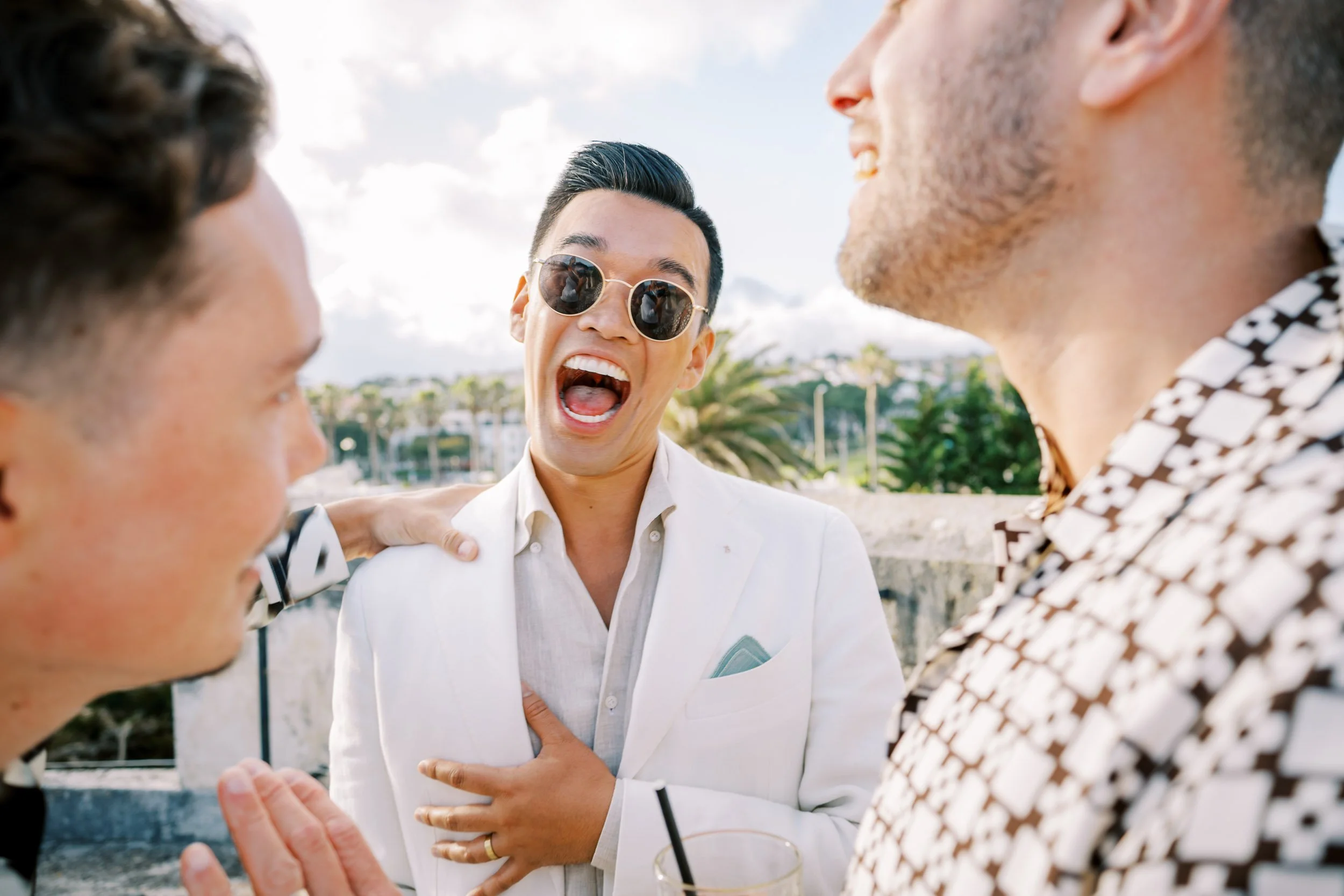 Three wedding guests laughing and chatting outdoors on a sunny day, with palm trees in the background. One man in the center is wearing sunglasses and a white blazer, holding his chest, and appears to be very happy.