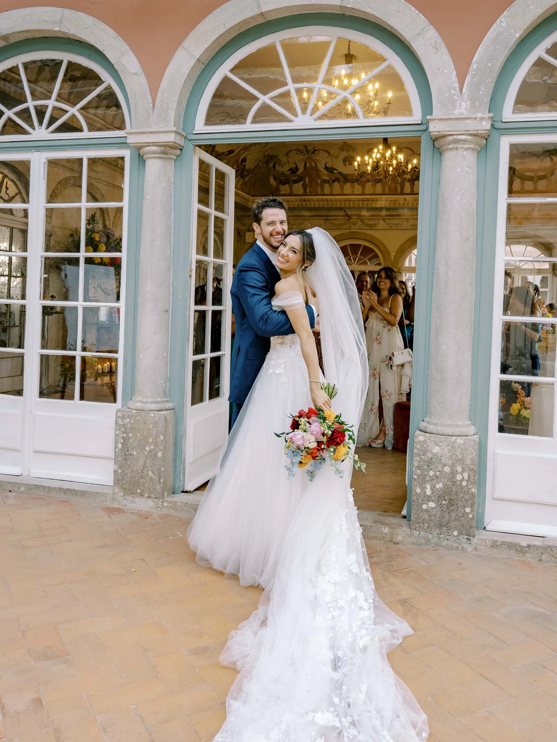 A bride and groom embracing at their wedding entrance, with guests in the background inside Casa dos Penedos in Sintra.