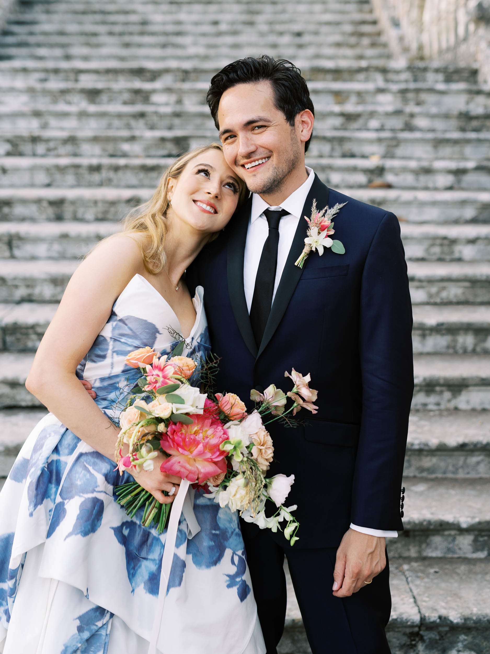 A bride and groom standing on outdoor steps, smiling and embracing each other. The bride holds a colorful bouquet, and the groom wears a dark suit with a boutonniere.