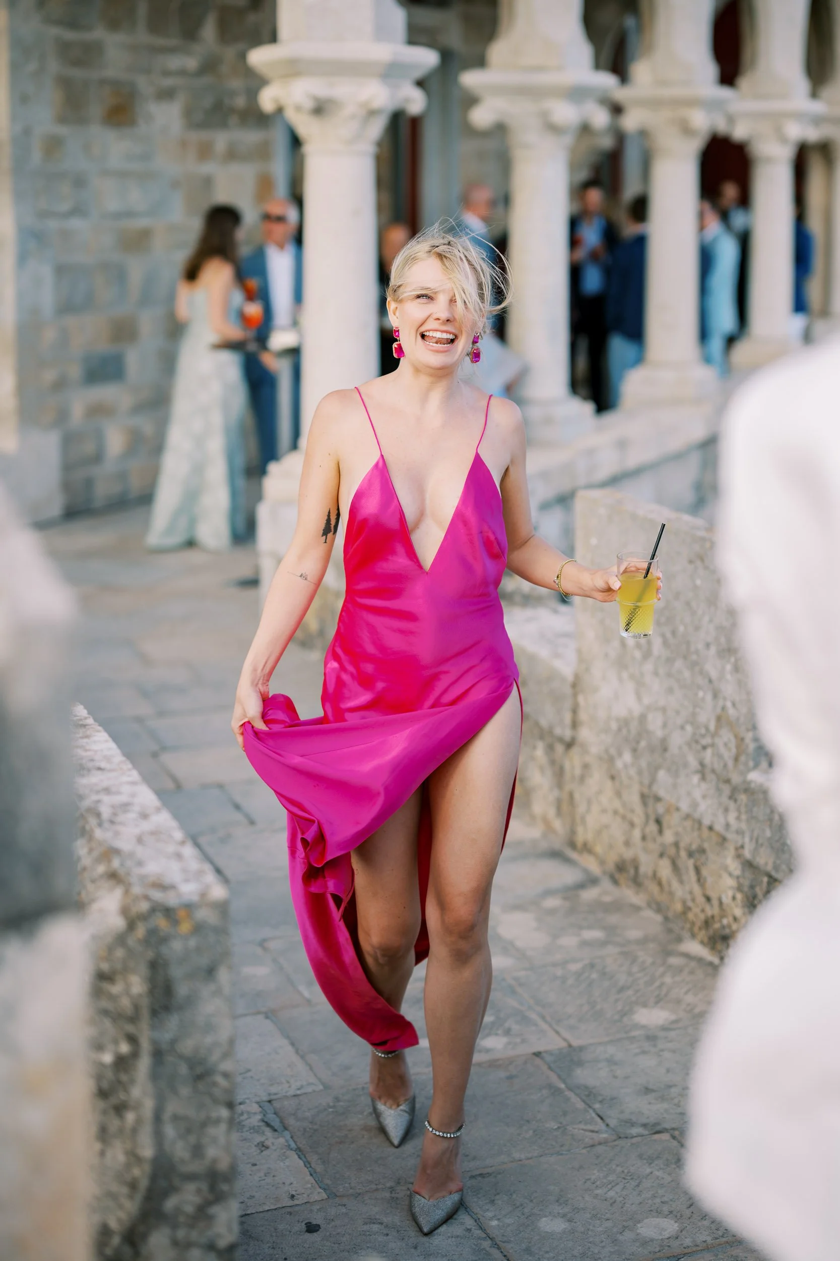 A wedding guest in a bright pink dress and high heels holding a drink, walking outdoors near stone architecture, with a happy expression at Forte da Cruz in Cascais, Portugal