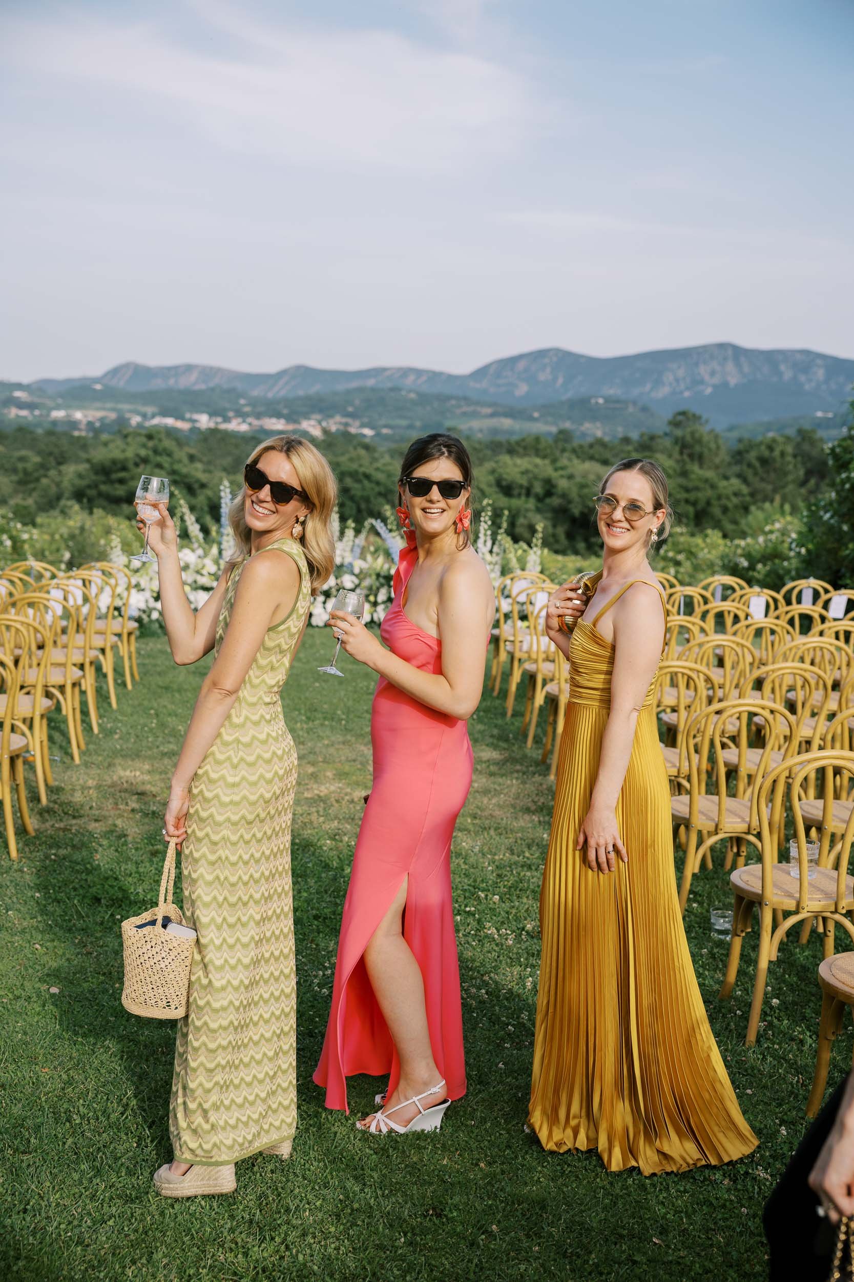 Three wedding guests in colorful dresses holding glasses at an outdoor event, with a backdrop of mountains and a clear sky at Herdade do Peru in Portugal.