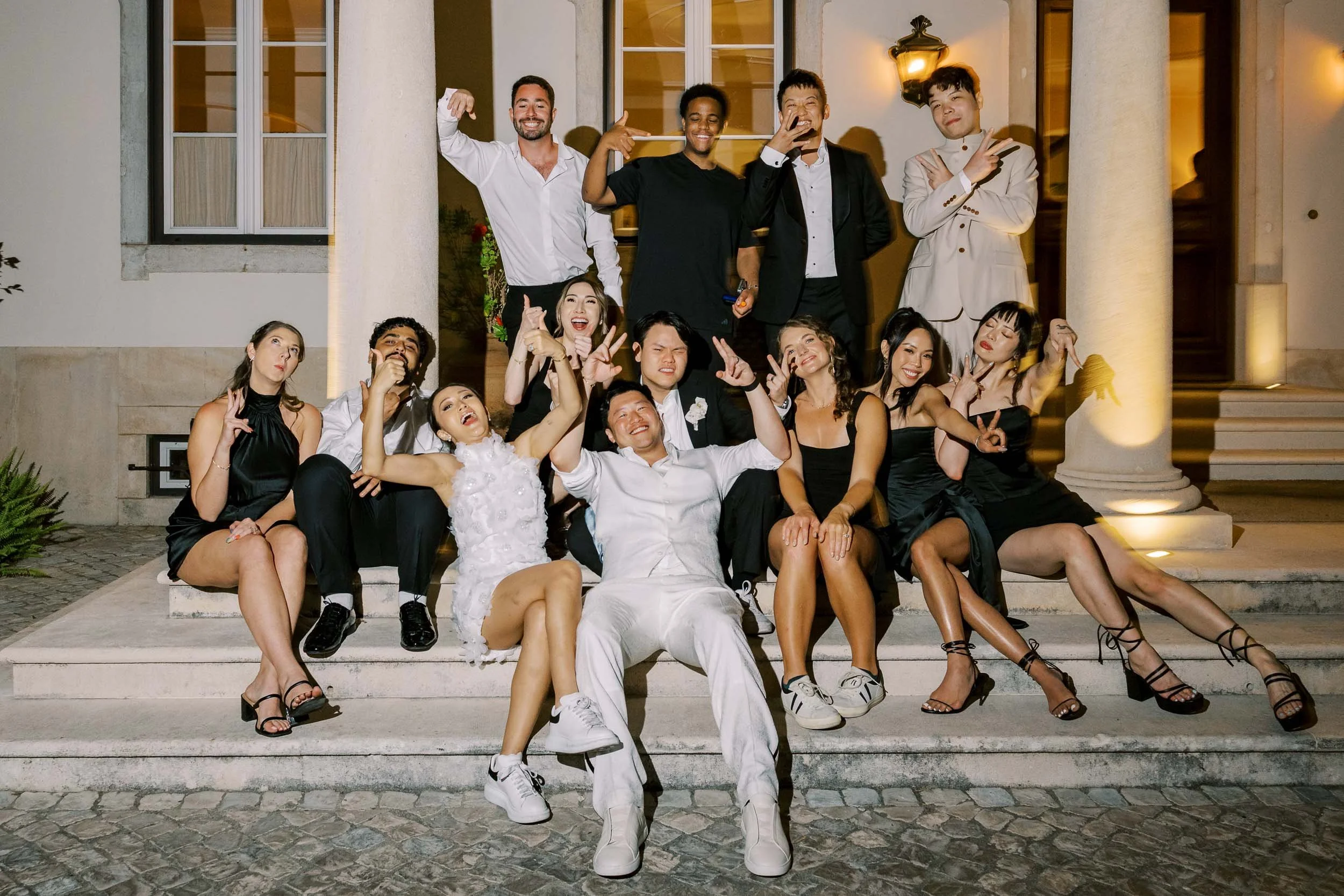 Group of wedding party celebrating on the steps of a Quinta da Bella Vista Palace at night, dressed in party attire, smiling, making playful gestures, and some holding drinks.
