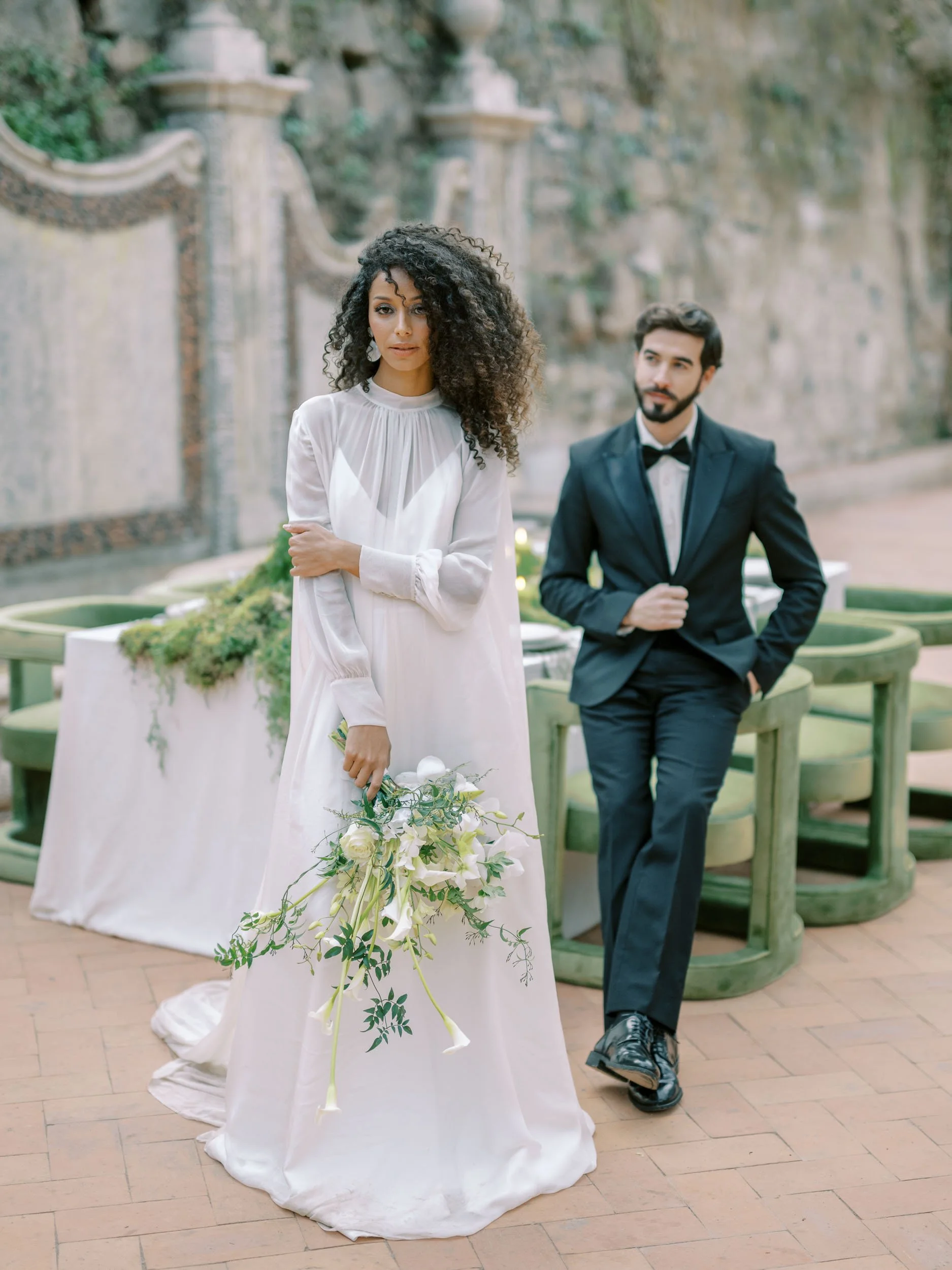 A woman in a white wedding dress holding a bouquet of white flowers, standing outdoors with a man in a black suit and bow tie in the background.