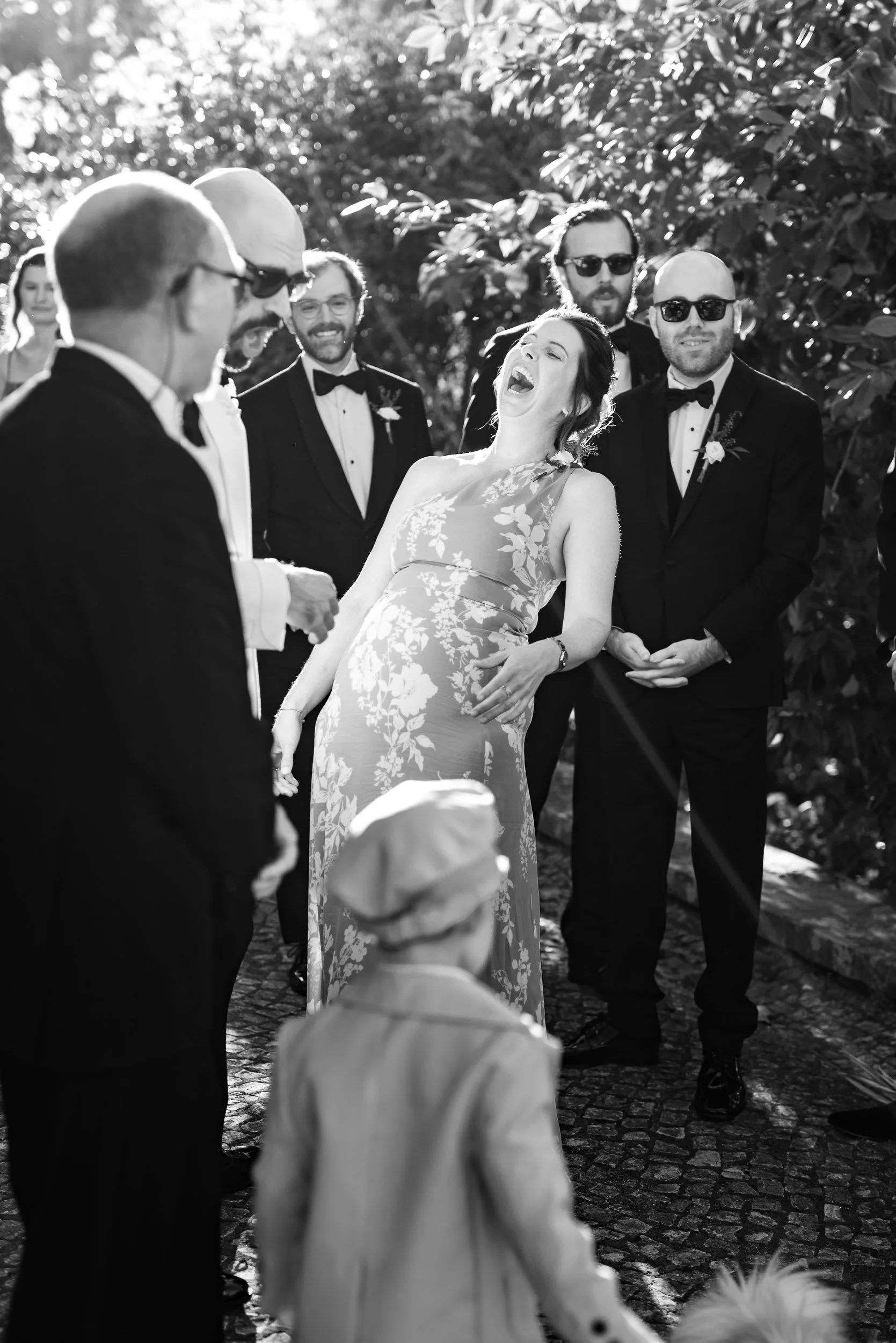 A wedding guest in a floral dress laughing among a group of men in tuxedos at an outdoor wedding.