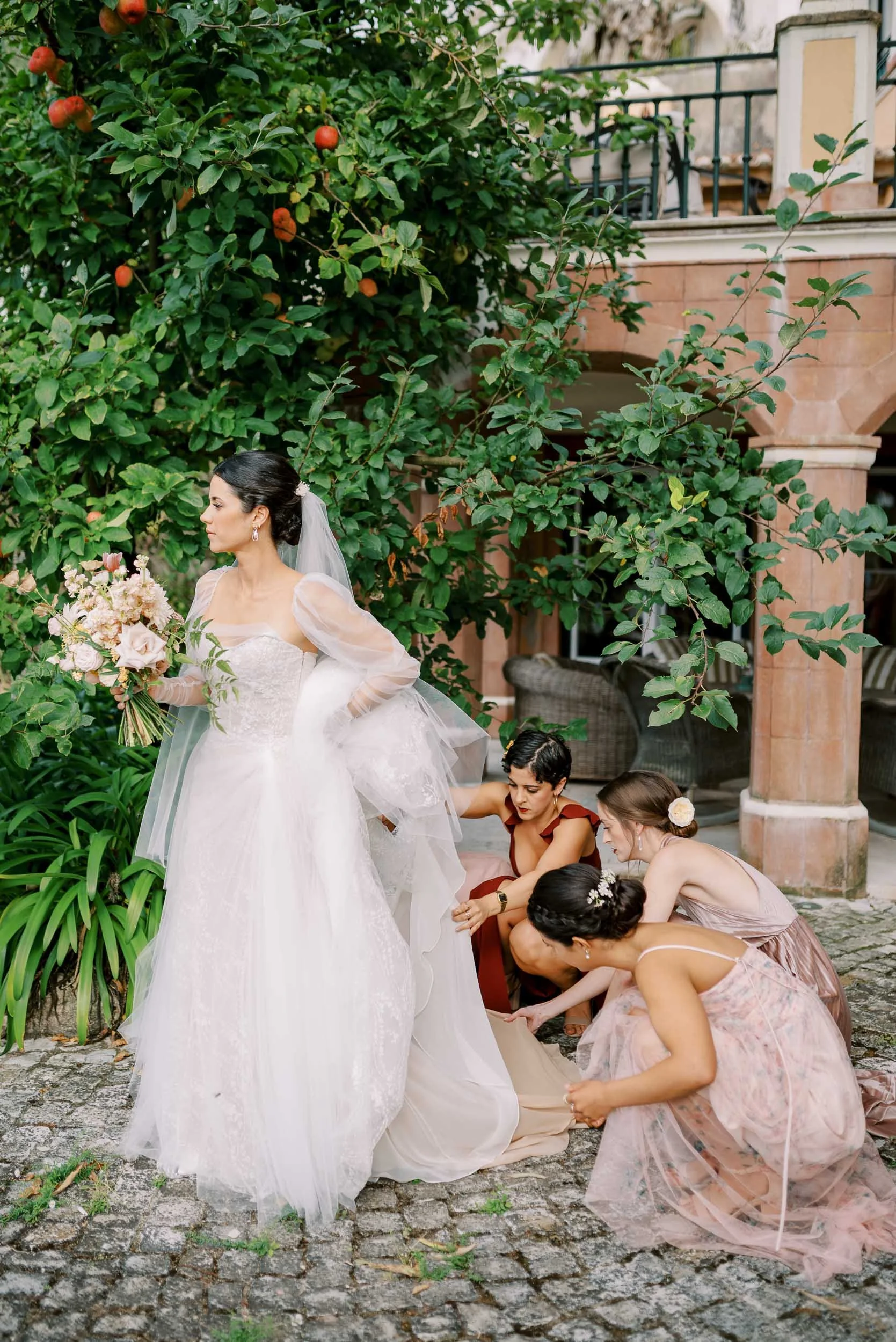 Bride in a white wedding gown holding a bouquet, four women adjusting her dress outdoors near a green bush and a building with stone columns.