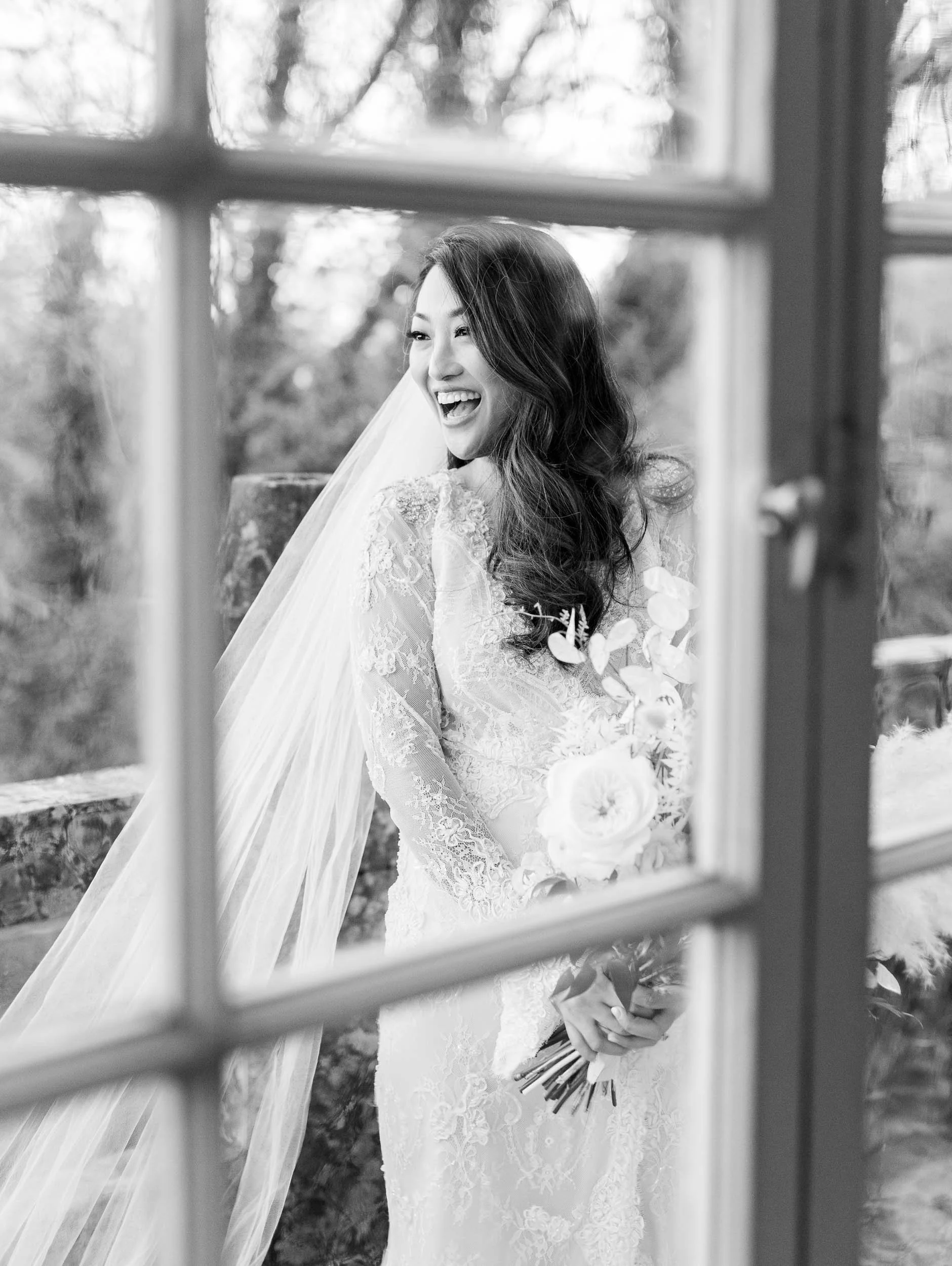 A bride with long, wavy hair wearing a lace wedding dress and veil, holding a bouquet of flowers, smiling and looking to the side, seen through a window with a blurred outdoor background.