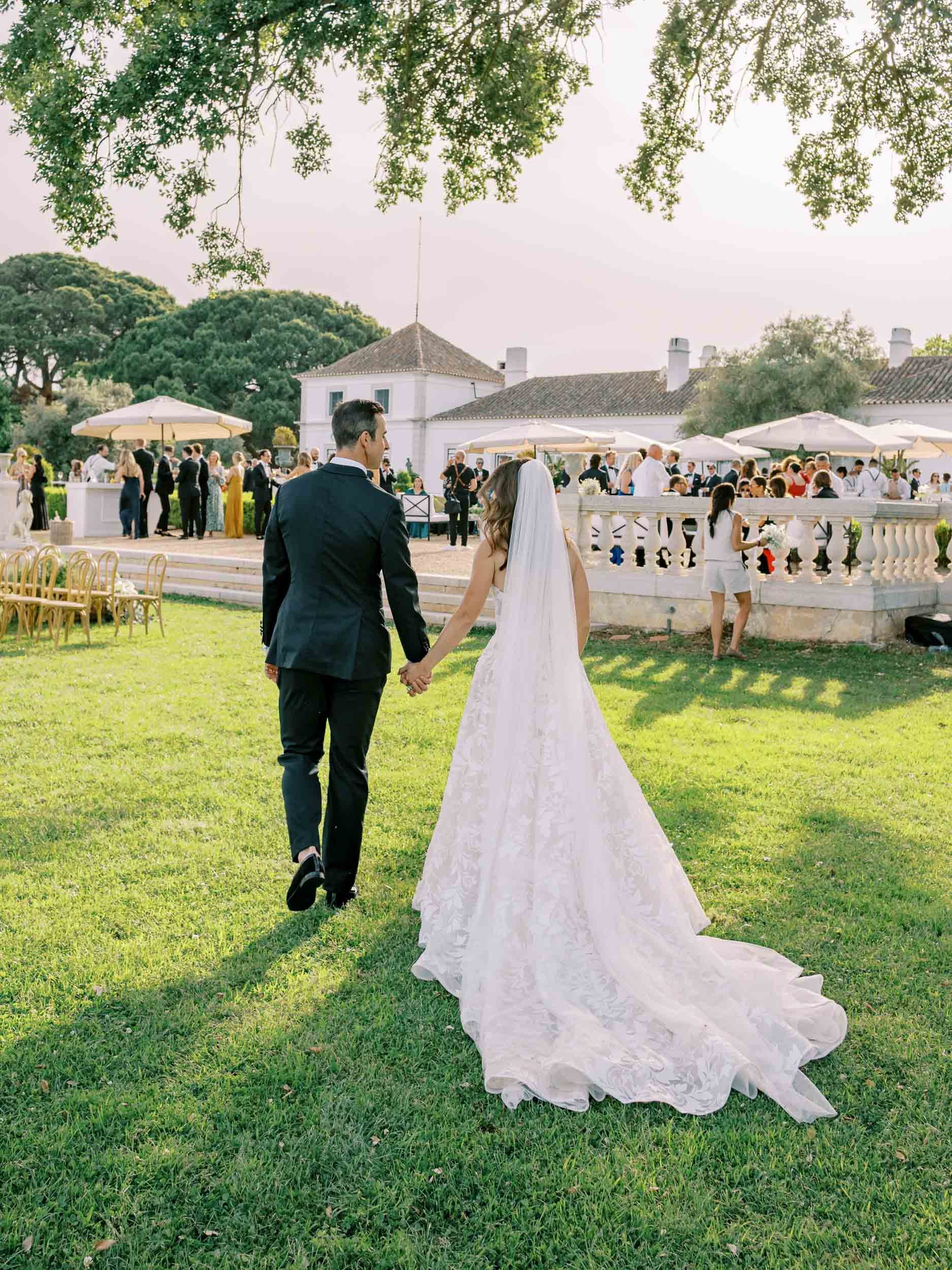 A bride and groom holding hands walking on a grassy lawn at an outdoor wedding reception, with guests in the background near white buildings and umbrellas, under large trees at Herdade do Perú in Portugal